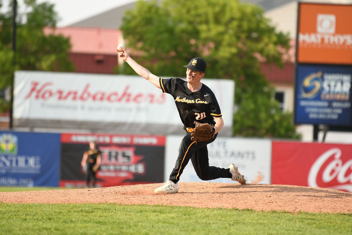 Pitcher in black uniform throws ball from mound with Coca-Cola advertisement visible in background.