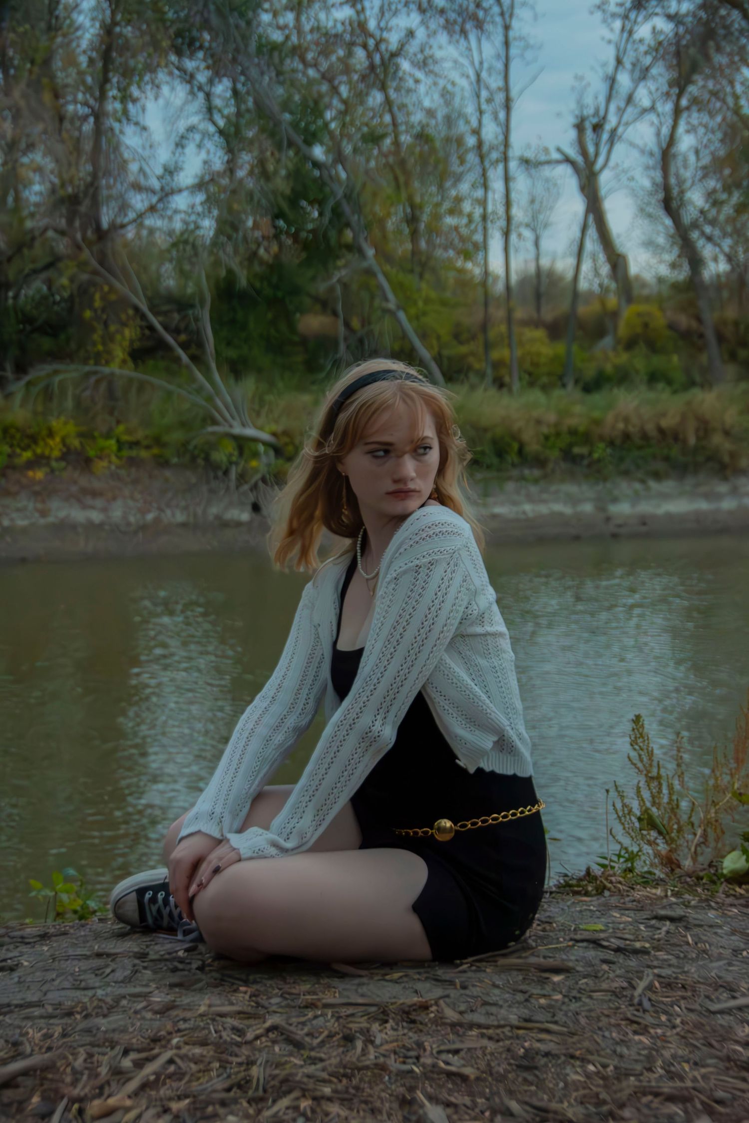 Person in white cardigan sitting by a riverbank during autumn.