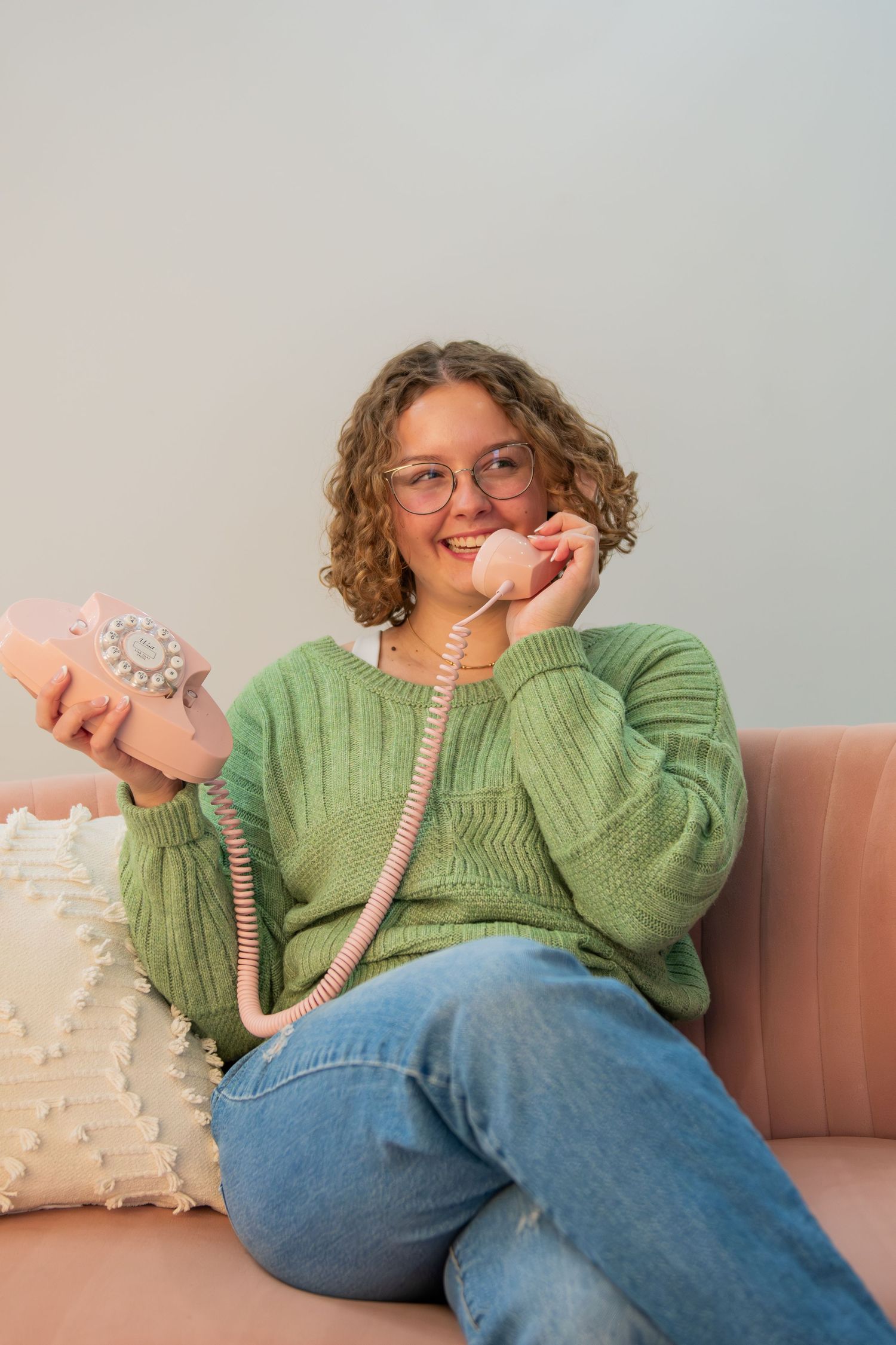 Person in green sweater and jeans lounges on beige couch while talking on pink phone with playful expressions.