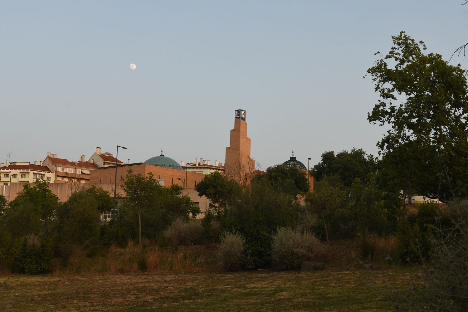 Silhouette of a town with prominent tower at sunset with moon visible in sky.