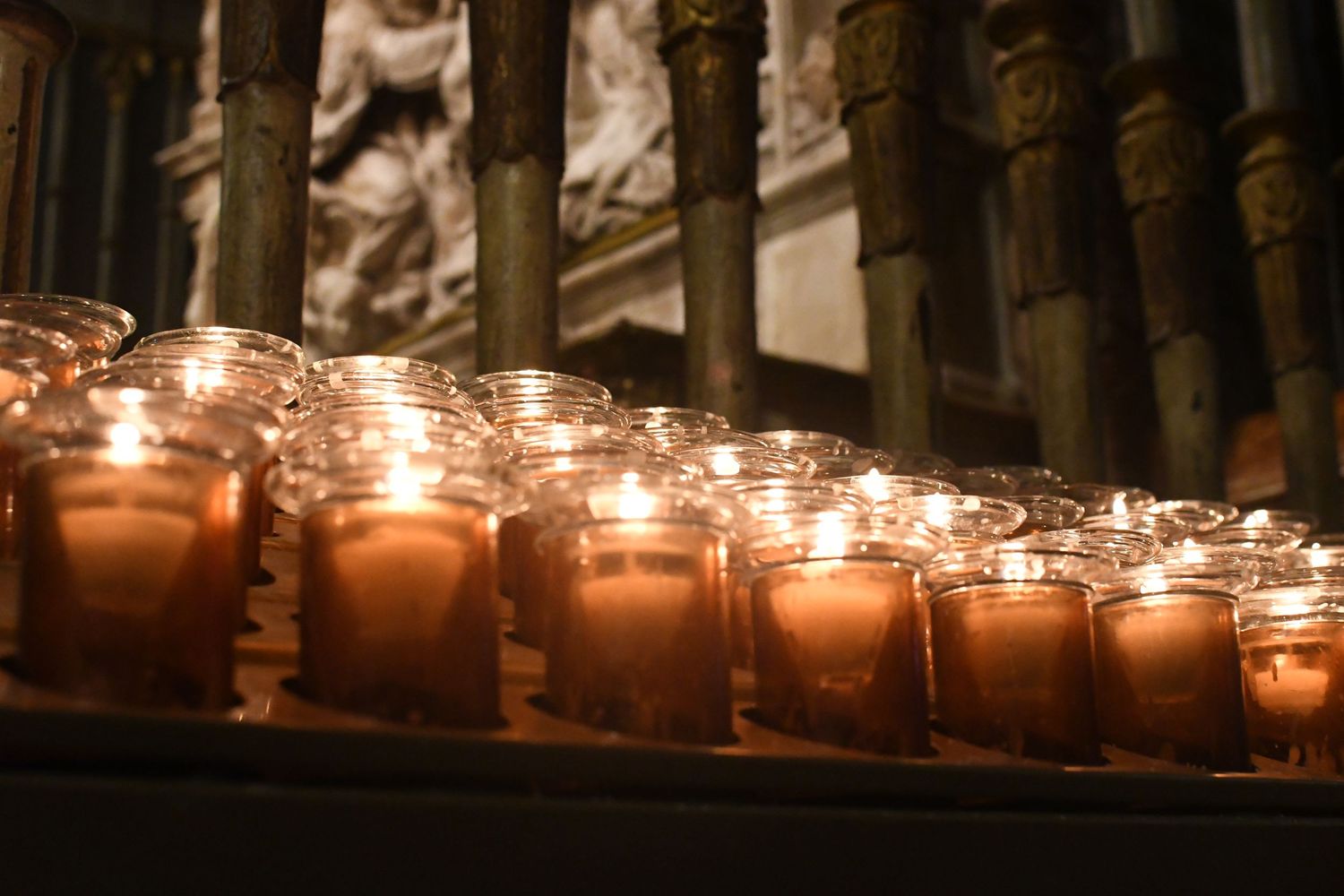 Row of glowing votive candles in glass holders inside a church or cathedral.