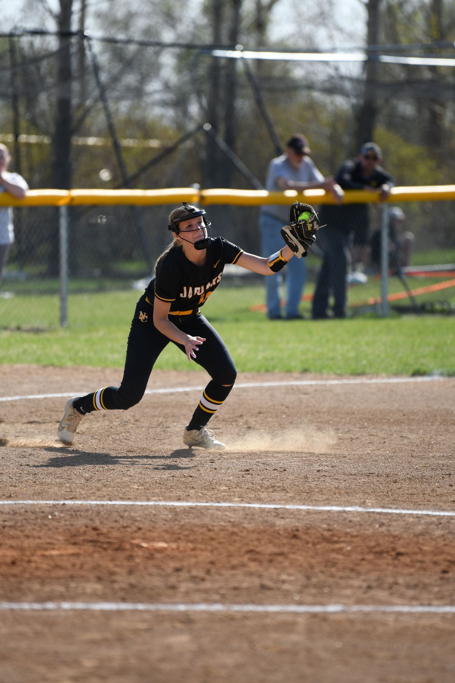 Player in black uniform dives and stretches for ball during softball game.
