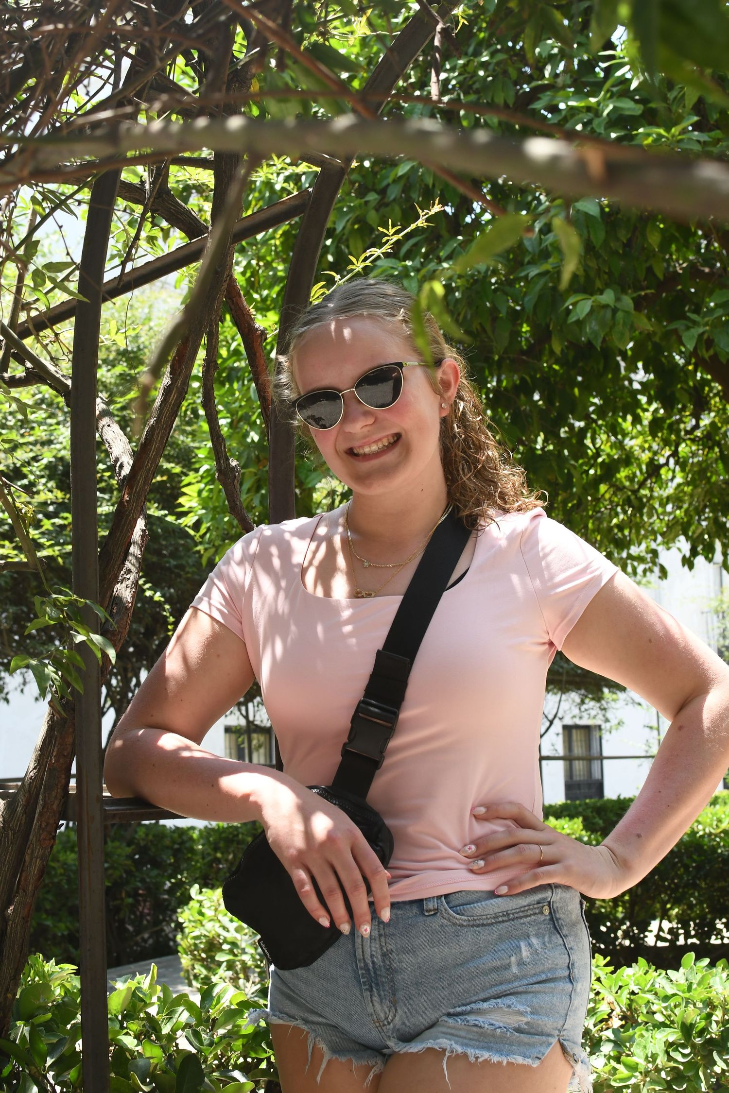 Tourist in pink shirt and denim shorts poses in sunny garden setting with lush greenery.