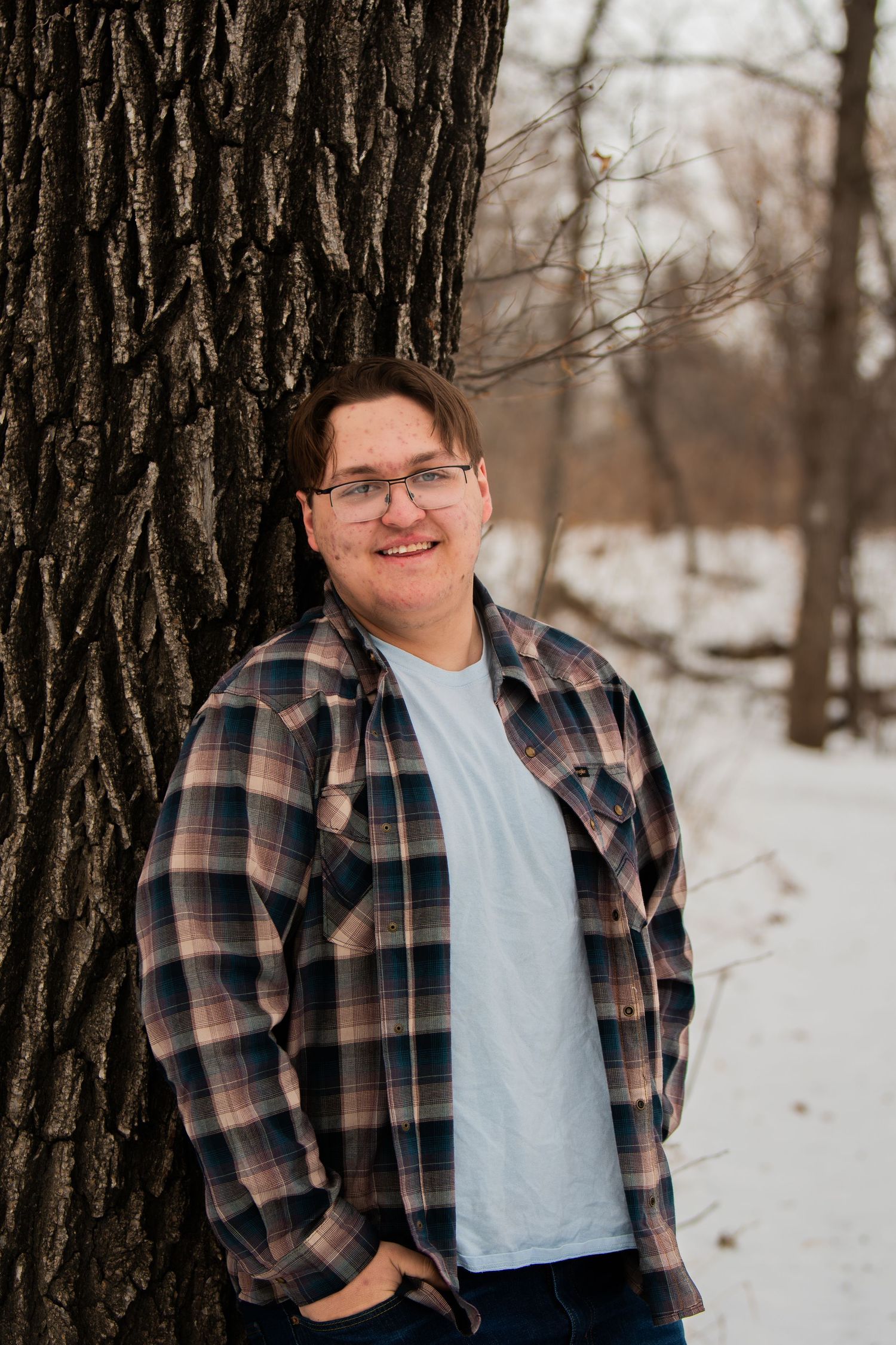 Person wearing plaid flannel shirt and light blue t-shirt poses against snowy winter forest background.