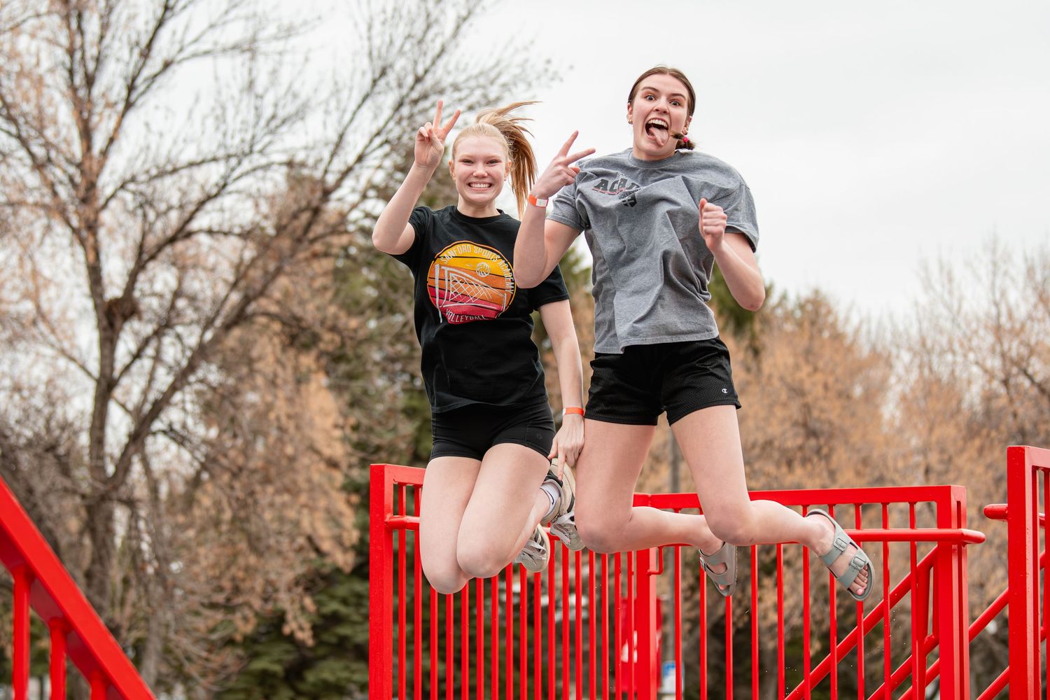Two young women jumping joyfully on a red bridge outdoors on a sunny day.
