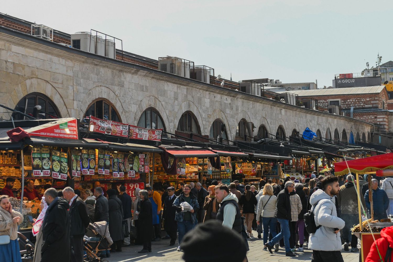 Panoramic view of a bustling outdoor market with vendor stalls and crowds under covered archways.