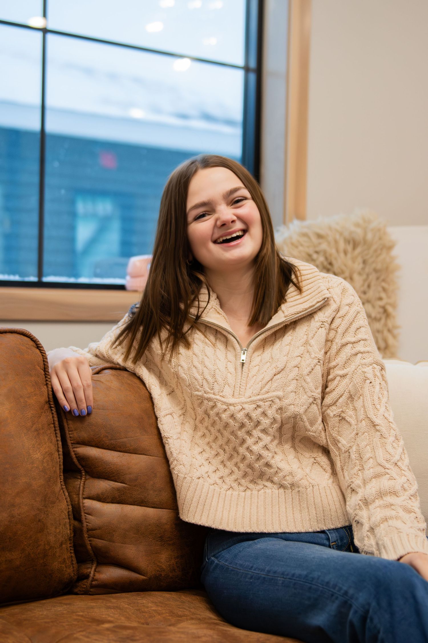 A person wearing a cream cable knit sweater sits comfortably on a brown leather couch near a window.