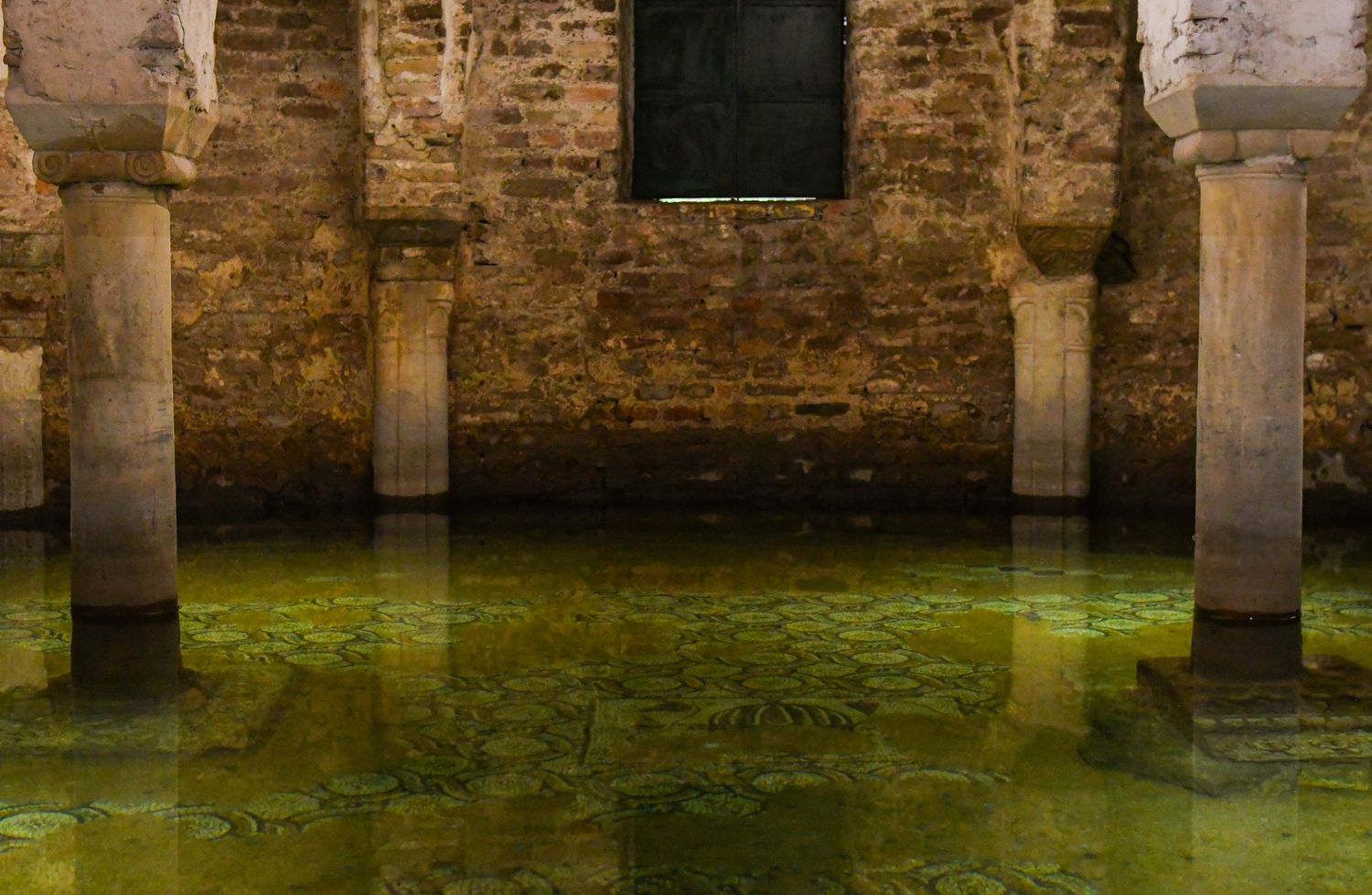 Ancient stone pillars rise from green water in a flooded underground chamber with weathered brick walls.
