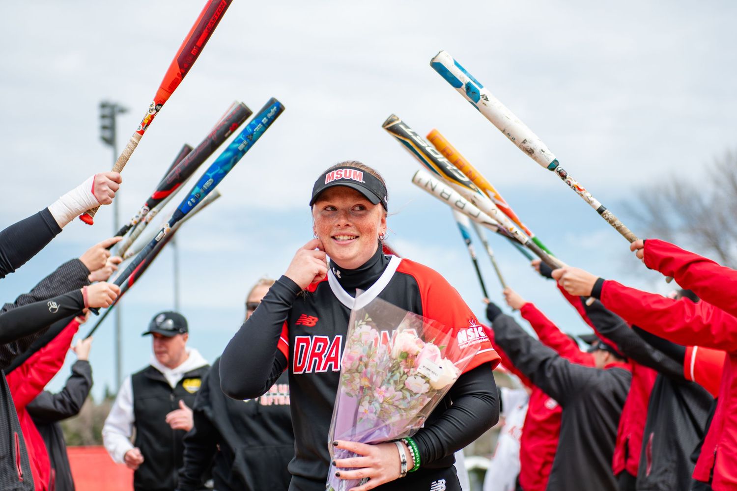 Softball player celebrated by teammates raising bats in arch, holding flowers, wearing Dragons jersey outdoors.