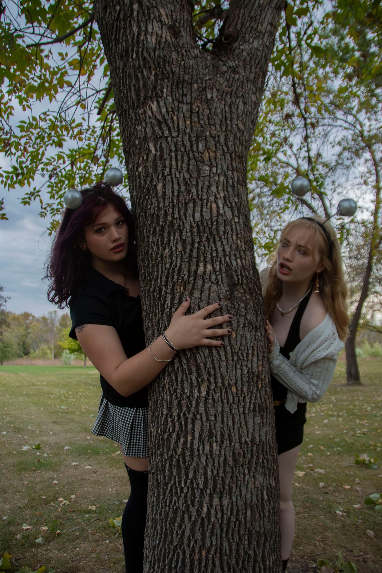 Two people playfully pose behind a large tree trunk in a park setting during autumn.