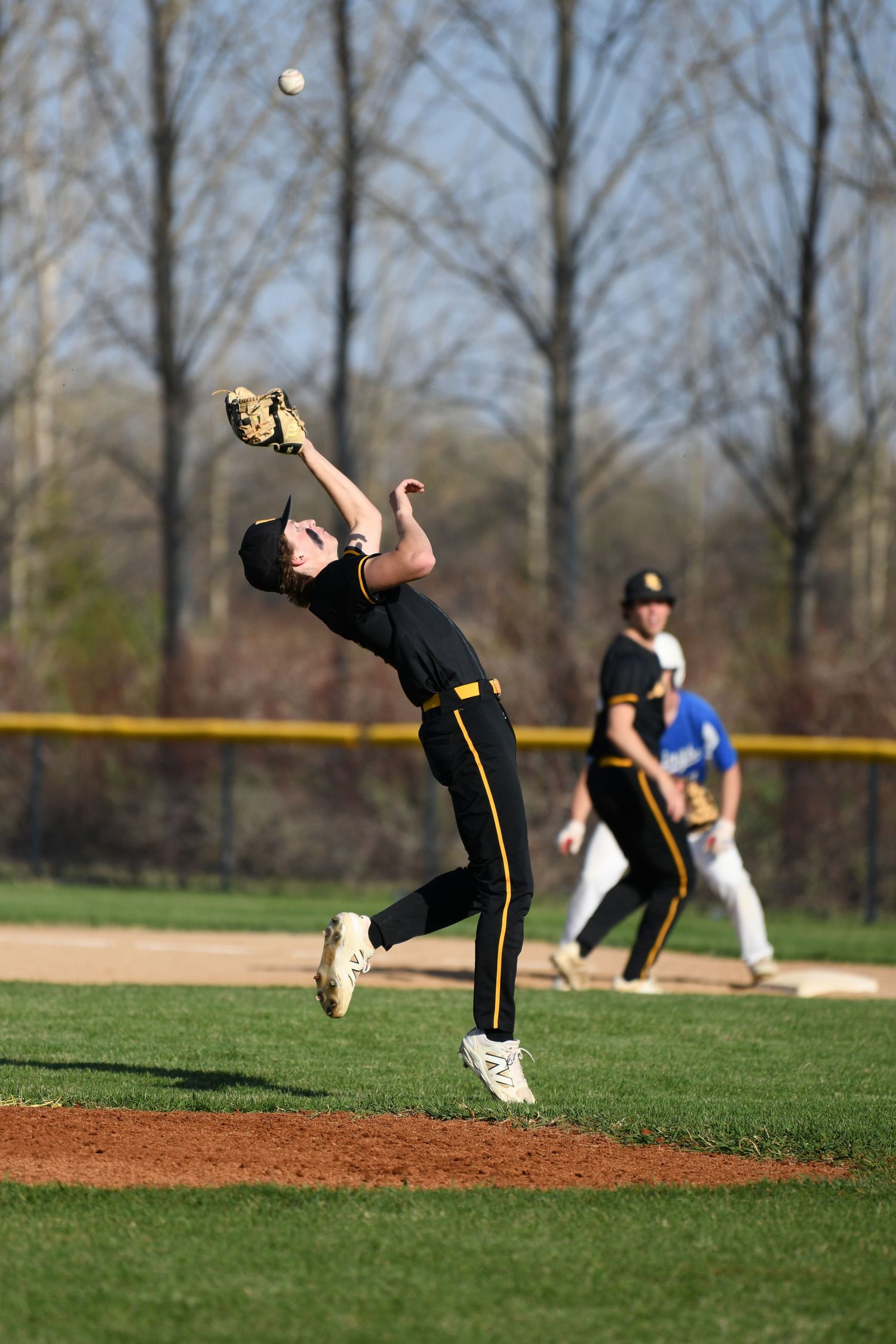 Baseball player leaps high to catch ball during game with trees in background.