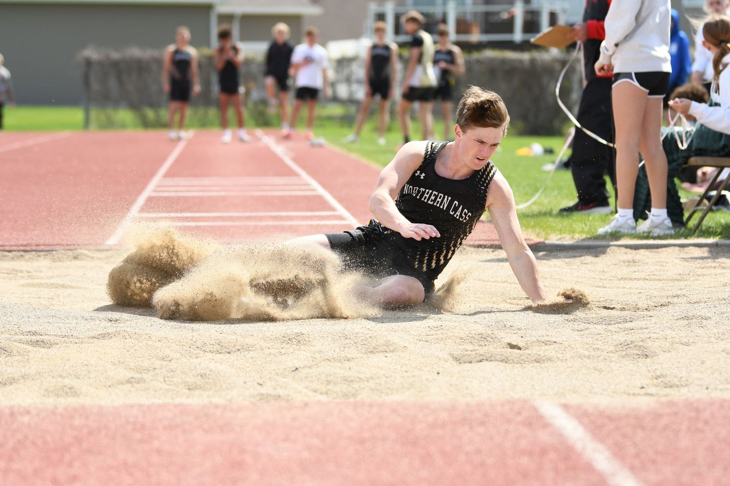 Track athlete lands in sand pit during long jump competition at outdoor meet.