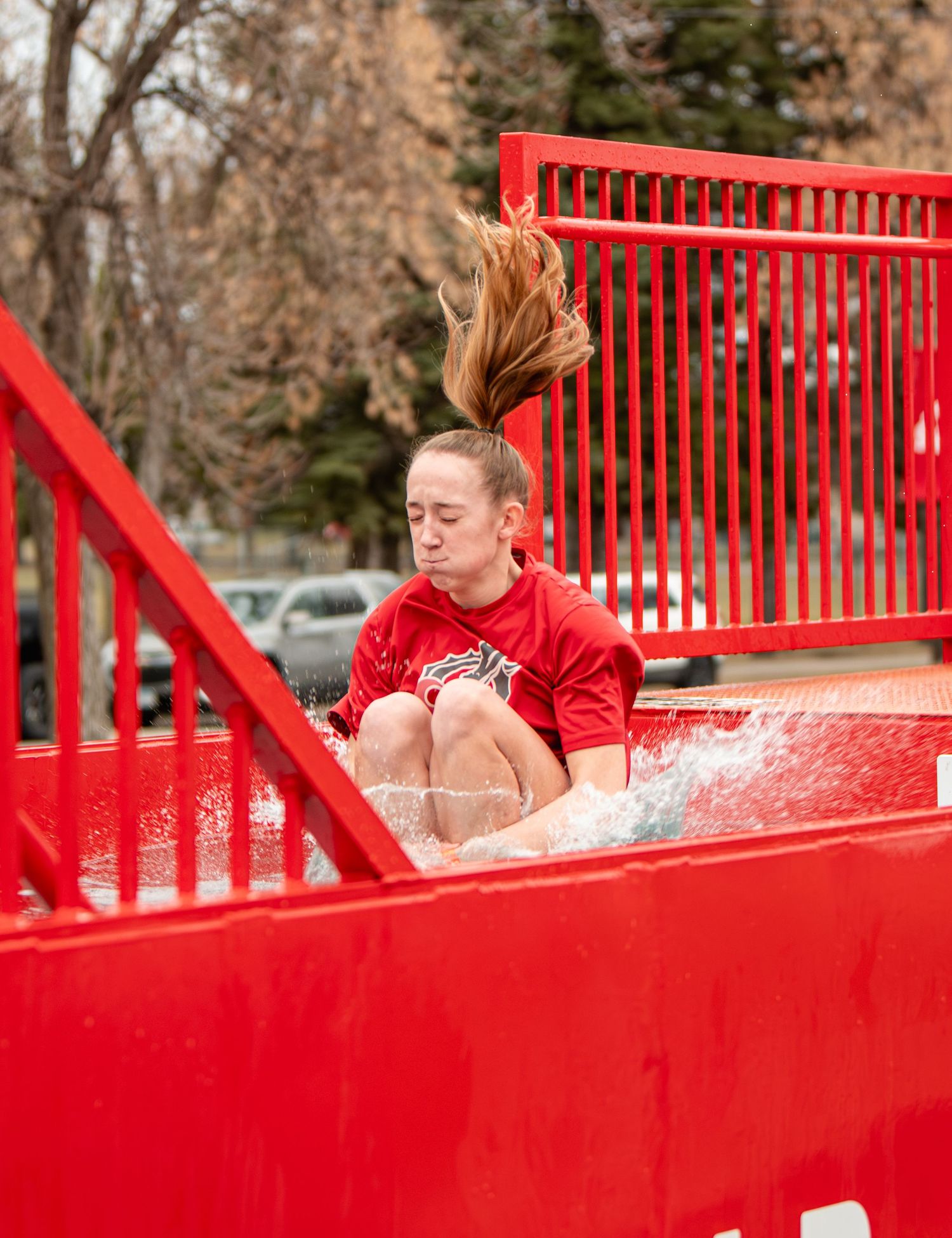 Woman in red shirt splashing into a dunk tank, hair flying, at an outdoor charity event.