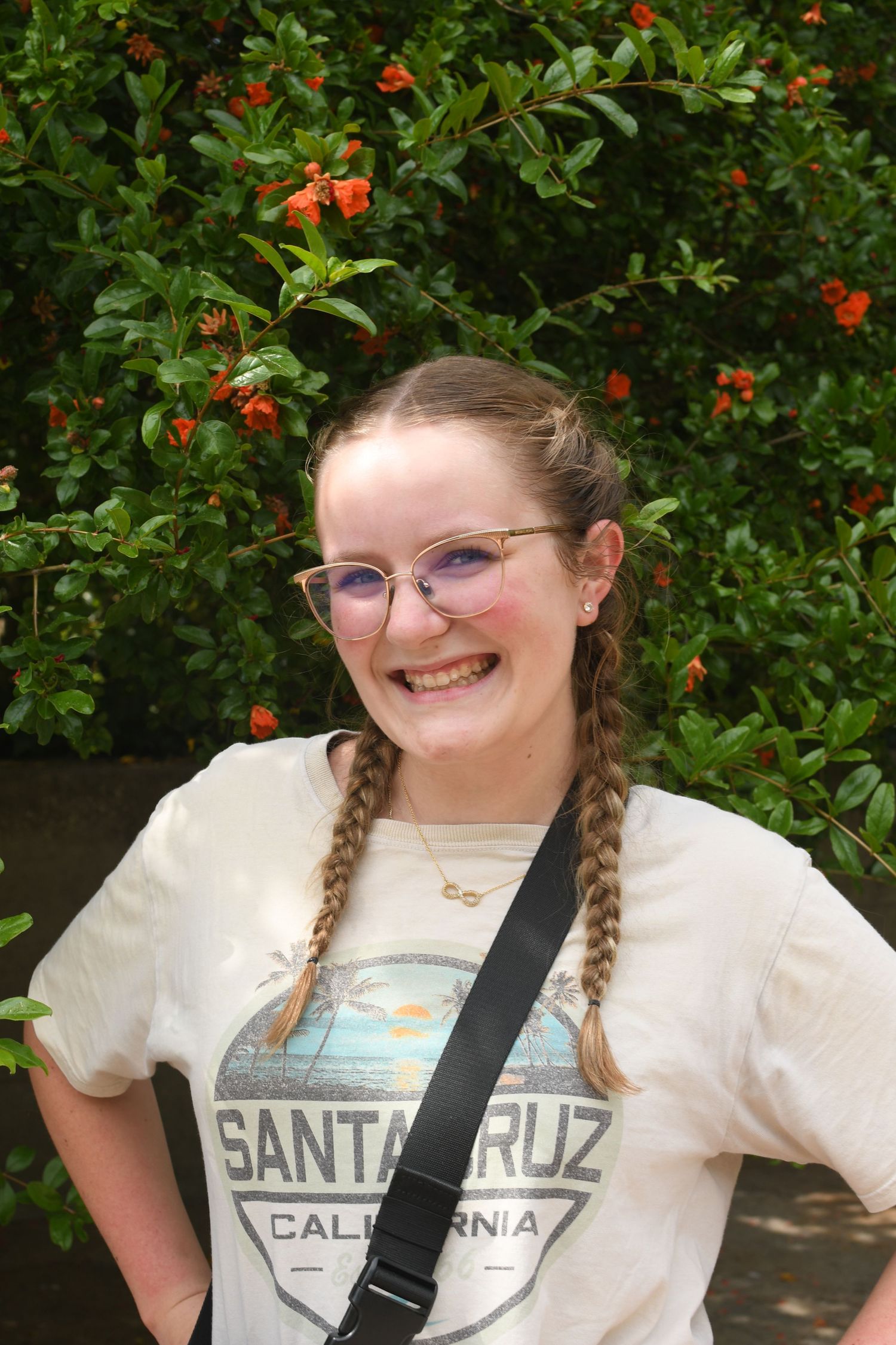 A smiling figure wearing glasses and a Santa Cruz t-shirt poses against a lush green backdrop with orange flowers.