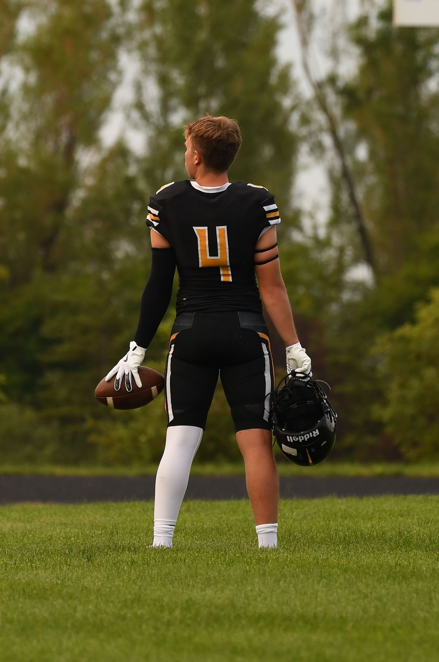 Football player in black uniform posing with helmet and ball on grass field.
