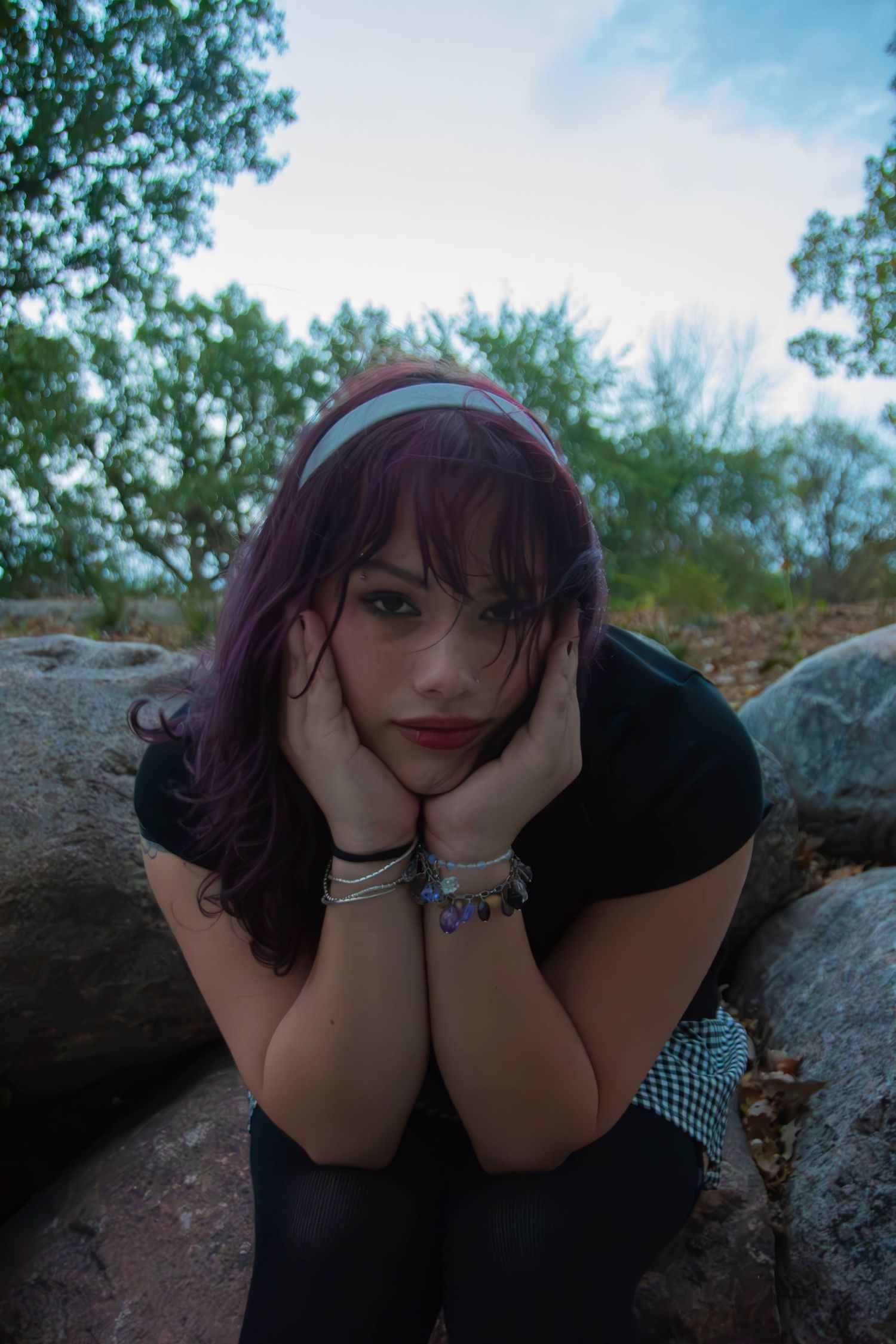 Person with purple hair and glasses resting their head on hands while lying on a rocky surface outdoors.