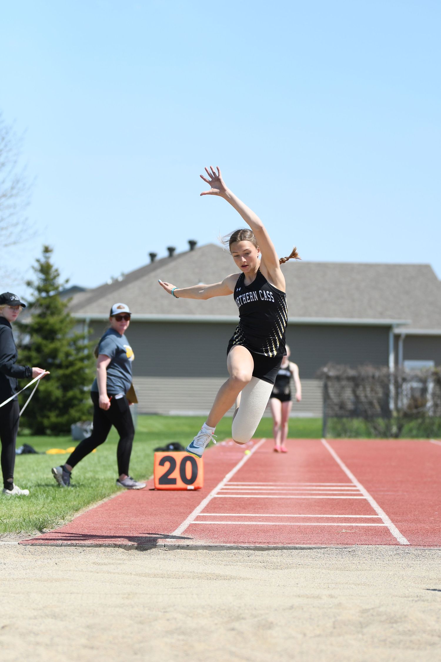 Athlete performs long jump at outdoor track meet with number 20 marker visible.