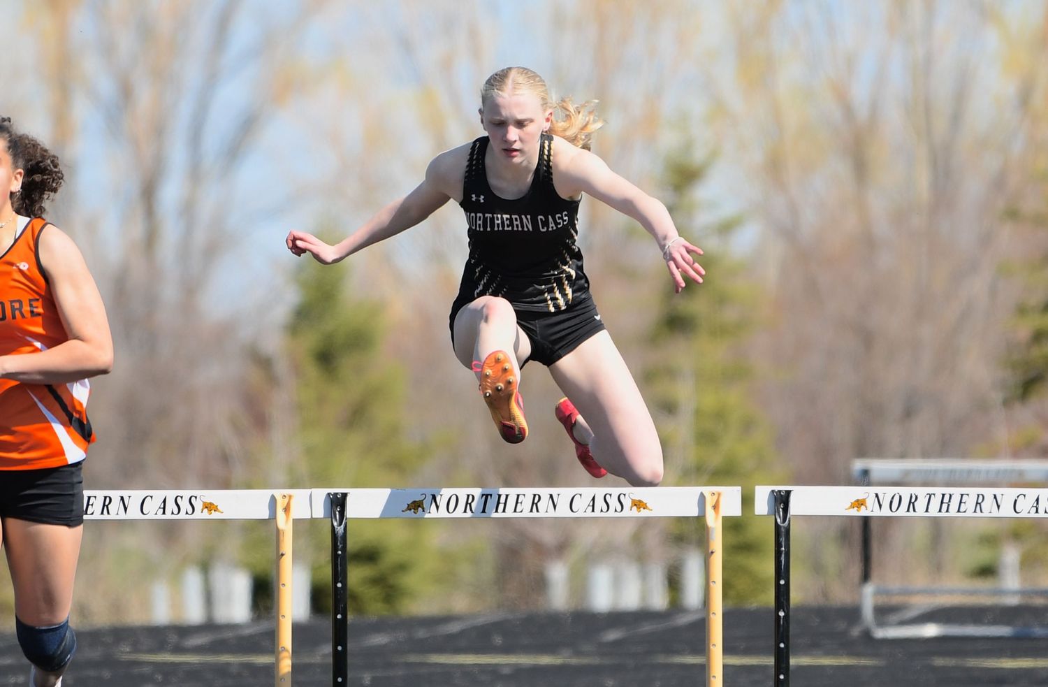 Athlete clearing a hurdle during track competition with Northern Cass branding visible.