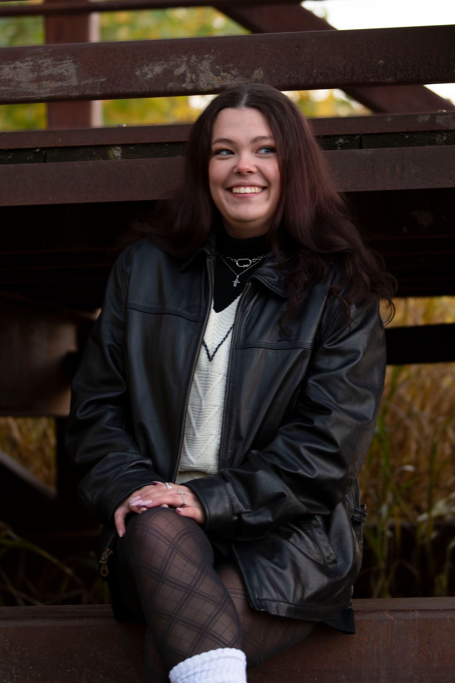 Person wearing black leather jacket and fishnet stockings sits on wooden bench in autumn setting.