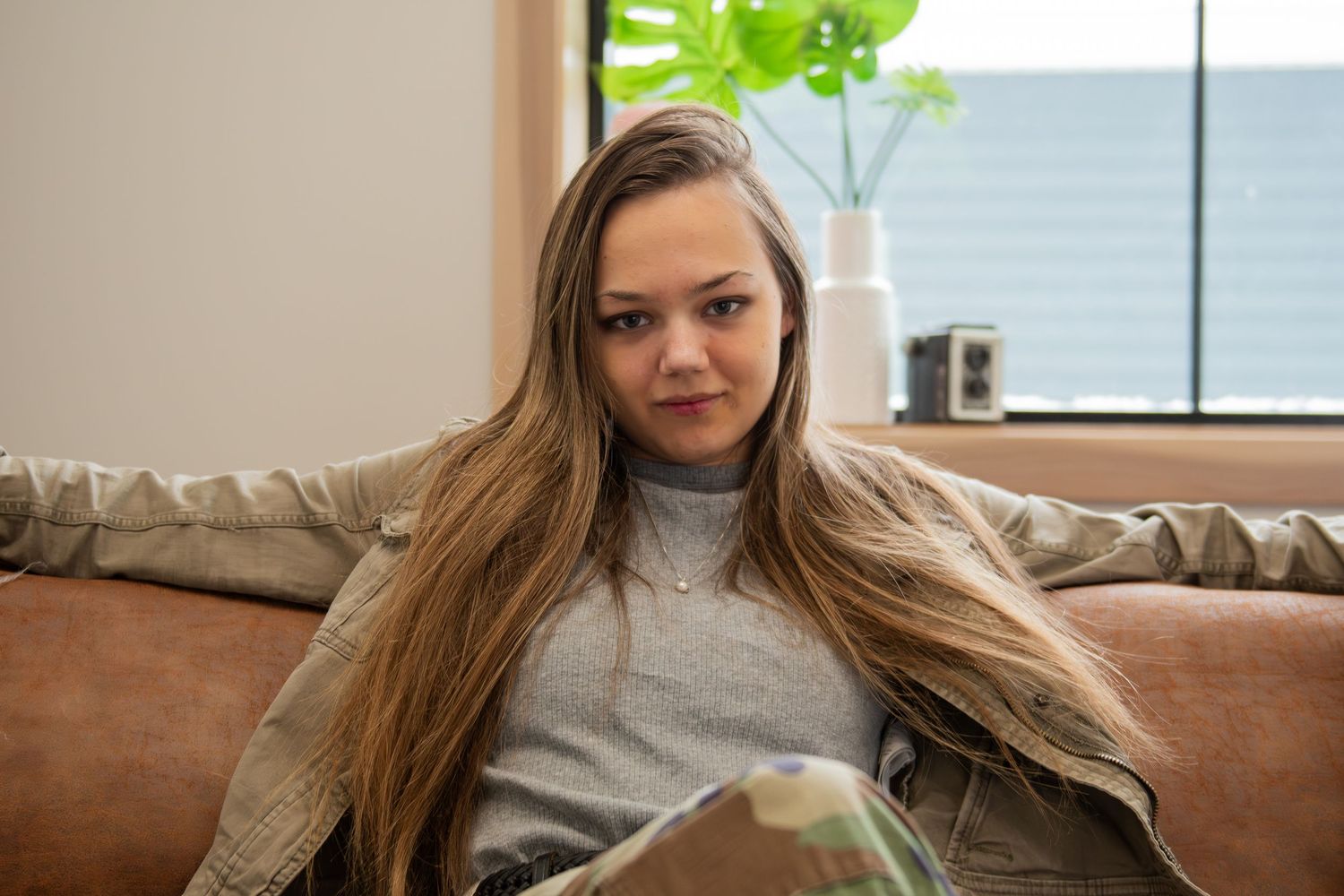 Woman with long hair sitting on couch at home wearing casual grey sweater near window with plants.