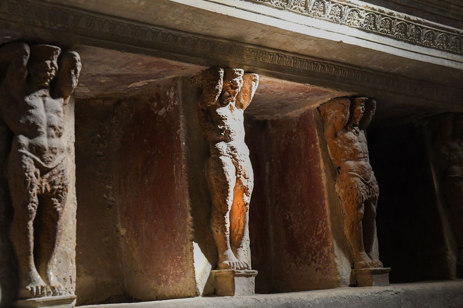 Row of ornate carved stone statues and columns in a dimly lit ancient corridor.