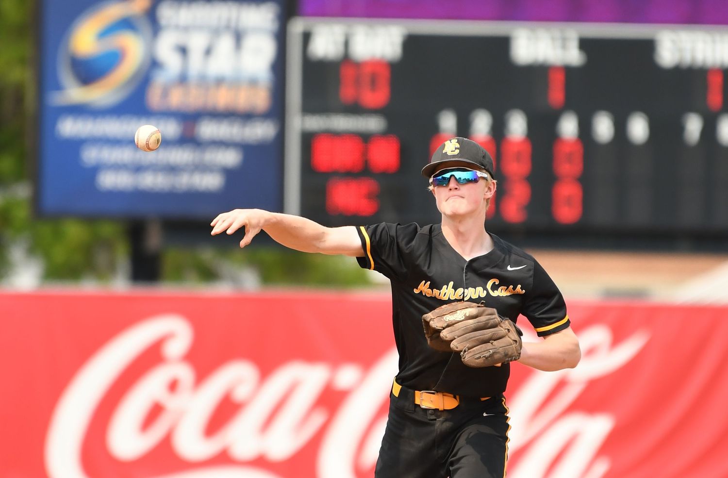 Baseball player in black uniform throws the ball with red Coca-Cola sign visible in background.