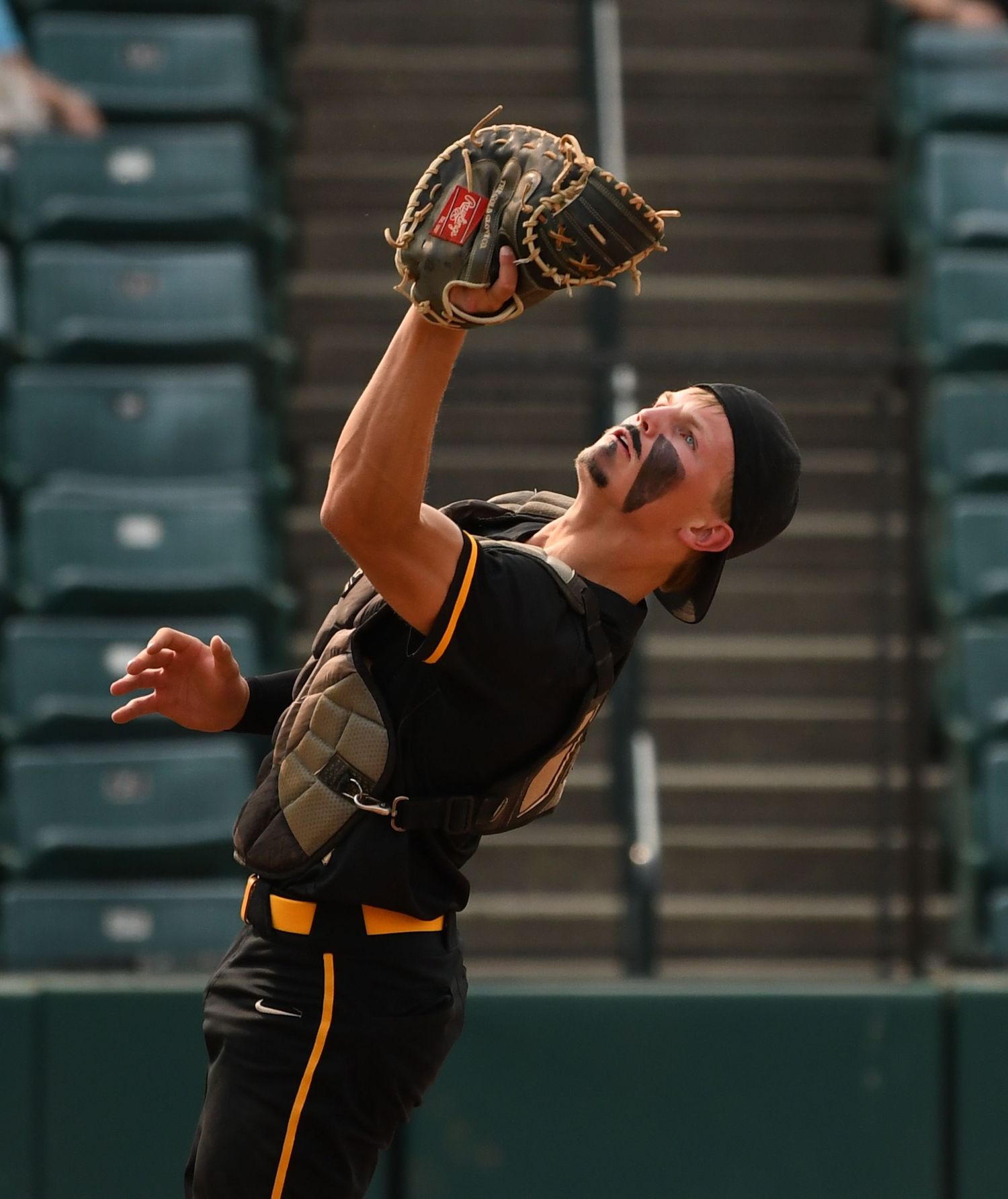 Baseball player in black uniform stretches to catch pop-up ball with stadium seats in background.