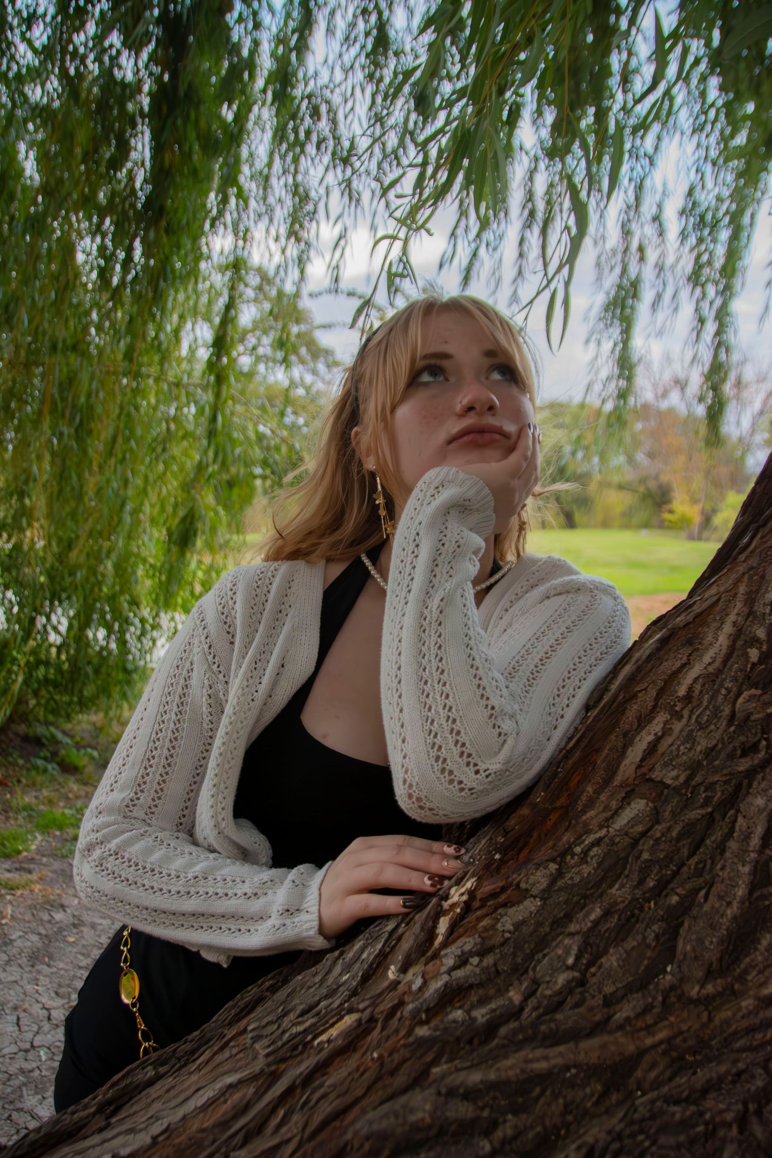 A person in a black top lies on a tree trunk under hanging willow branches in an outdoor setting.