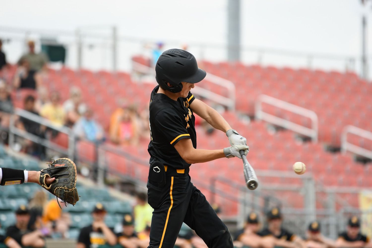 Baseball batter in black uniform swings the bat during a game with red stadium seats in background.