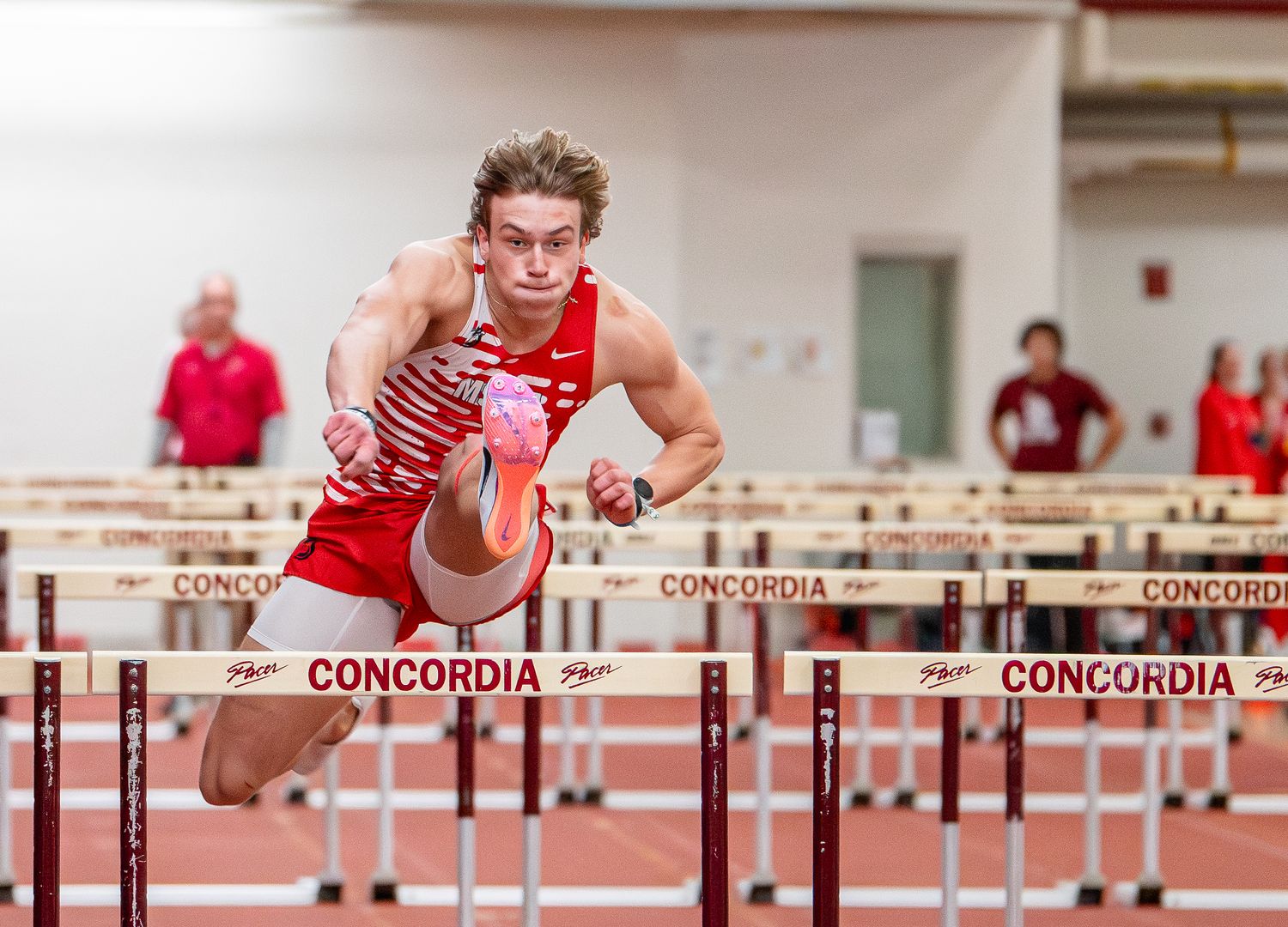 Male athlete in red uniform clears a hurdle during an indoor Concordia track and field competition.