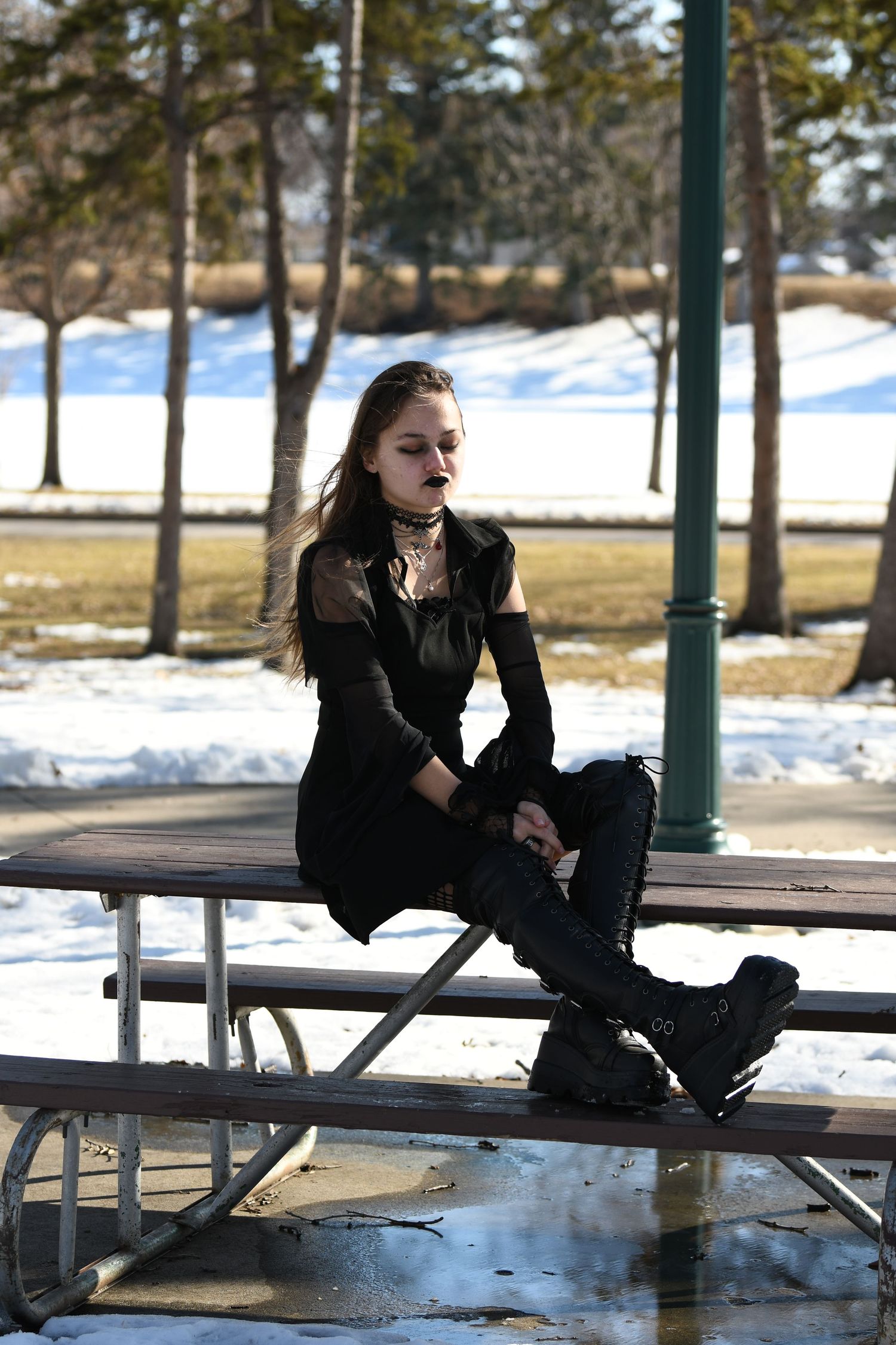 Person in dark clothing sits on a snowy picnic table in a winter park setting.