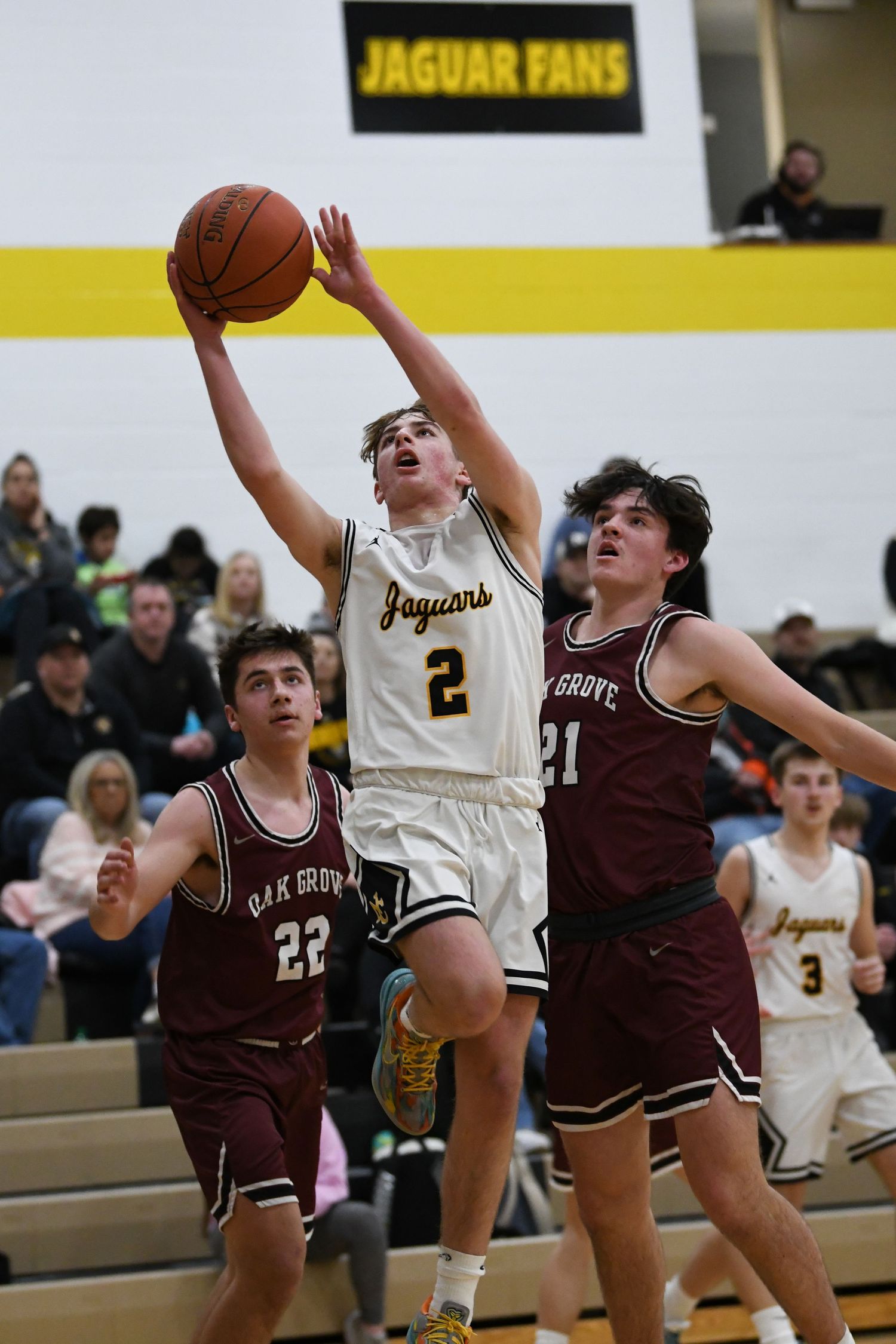 Basketball player in white jersey drives to the hoop between two defenders during intense game action.