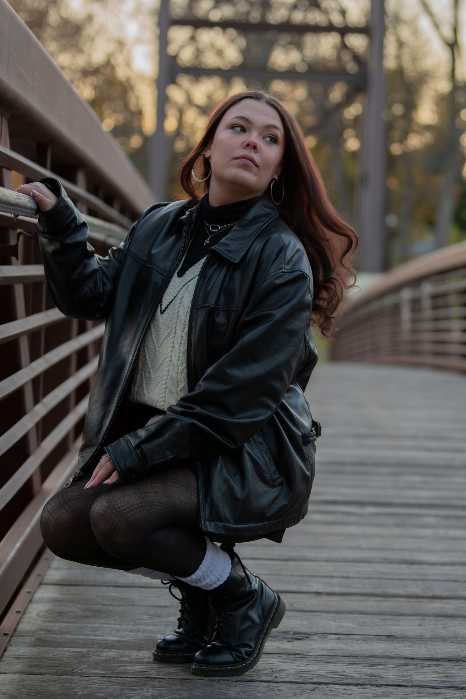A person in a black leather jacket and white sweater poses on a wooden boardwalk during golden hour.