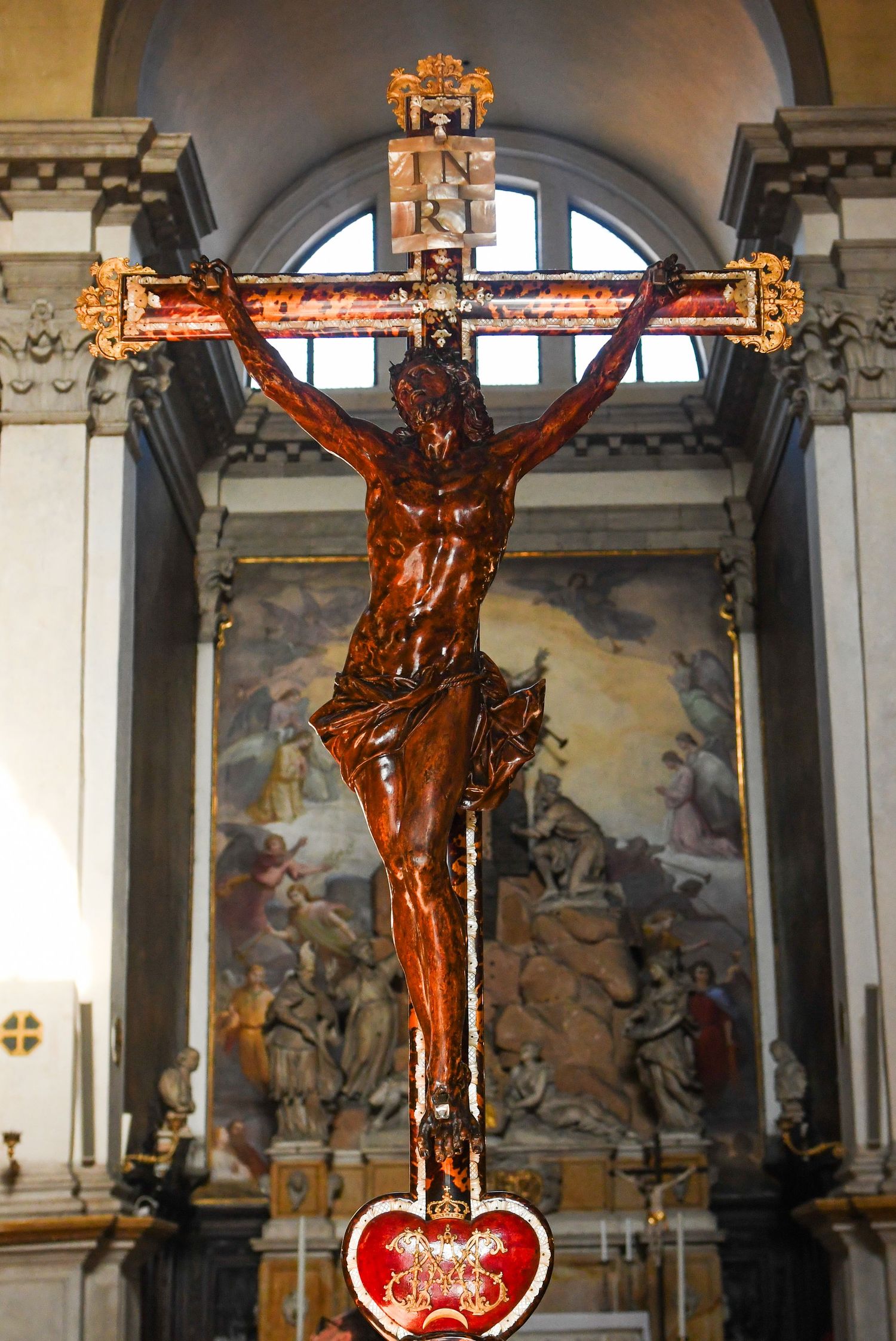 Bronze crucifix mounted on wall with religious artwork and murals in background.