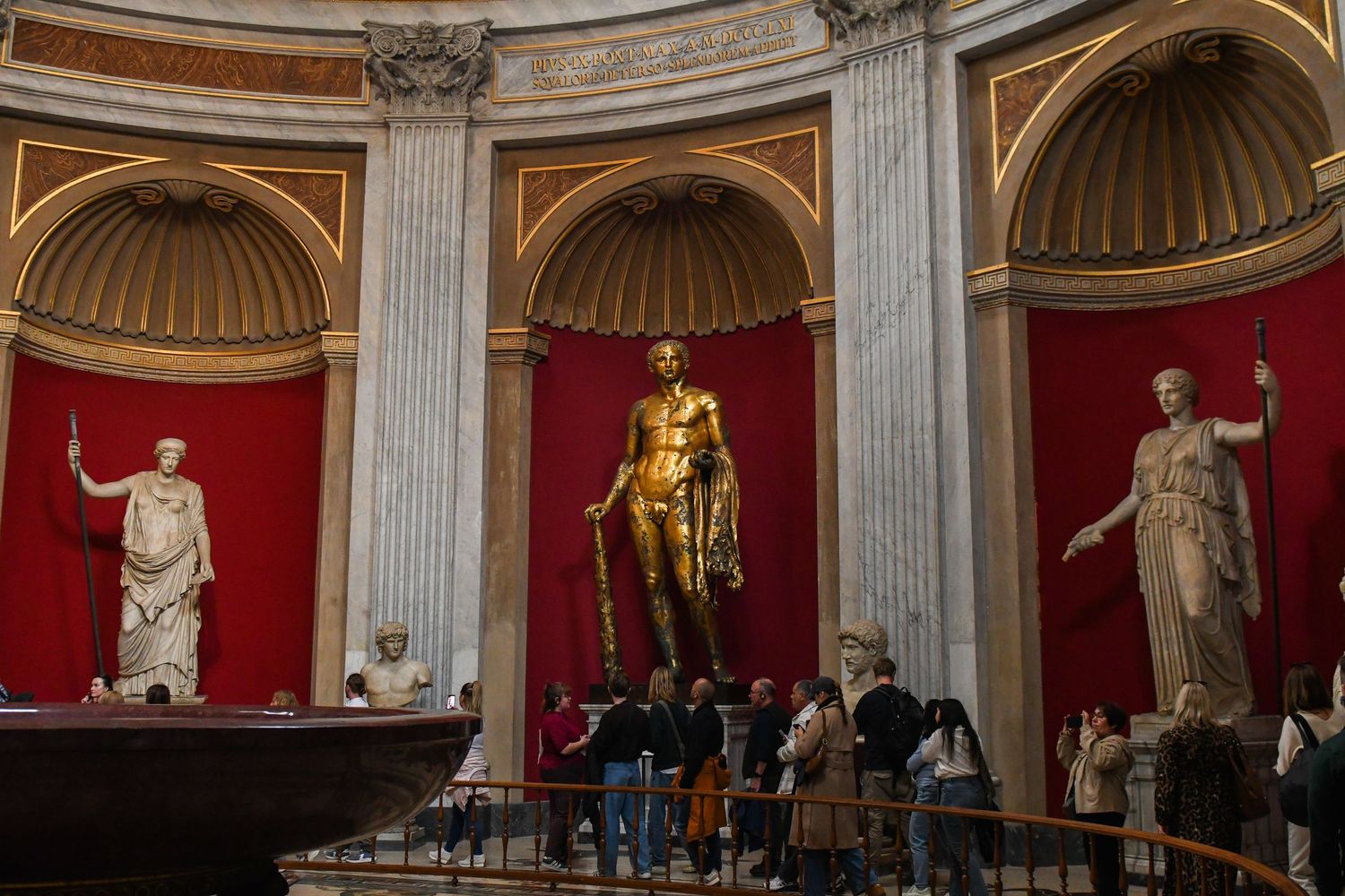Museum gallery with classical statues displayed against red walls in arched alcoves.