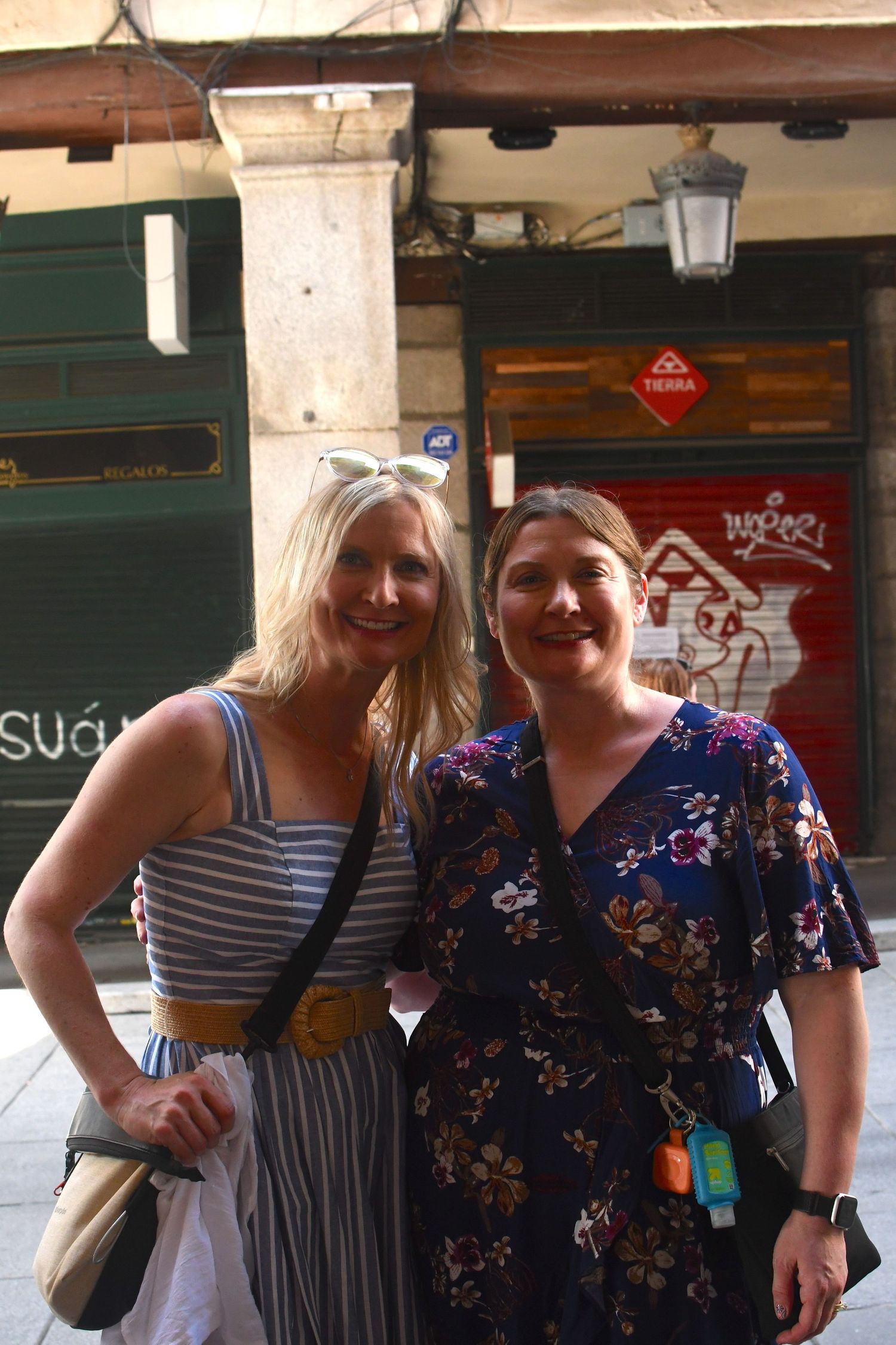 Two people posing together under stone archway near shops in European setting.