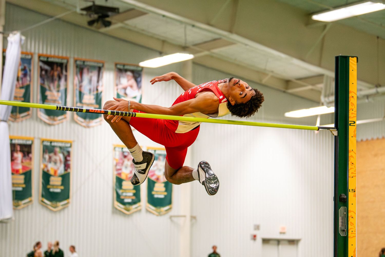 A gymnast performs a dynamic aerial flip in a bright gymnasium with green banners hanging from the ceiling.