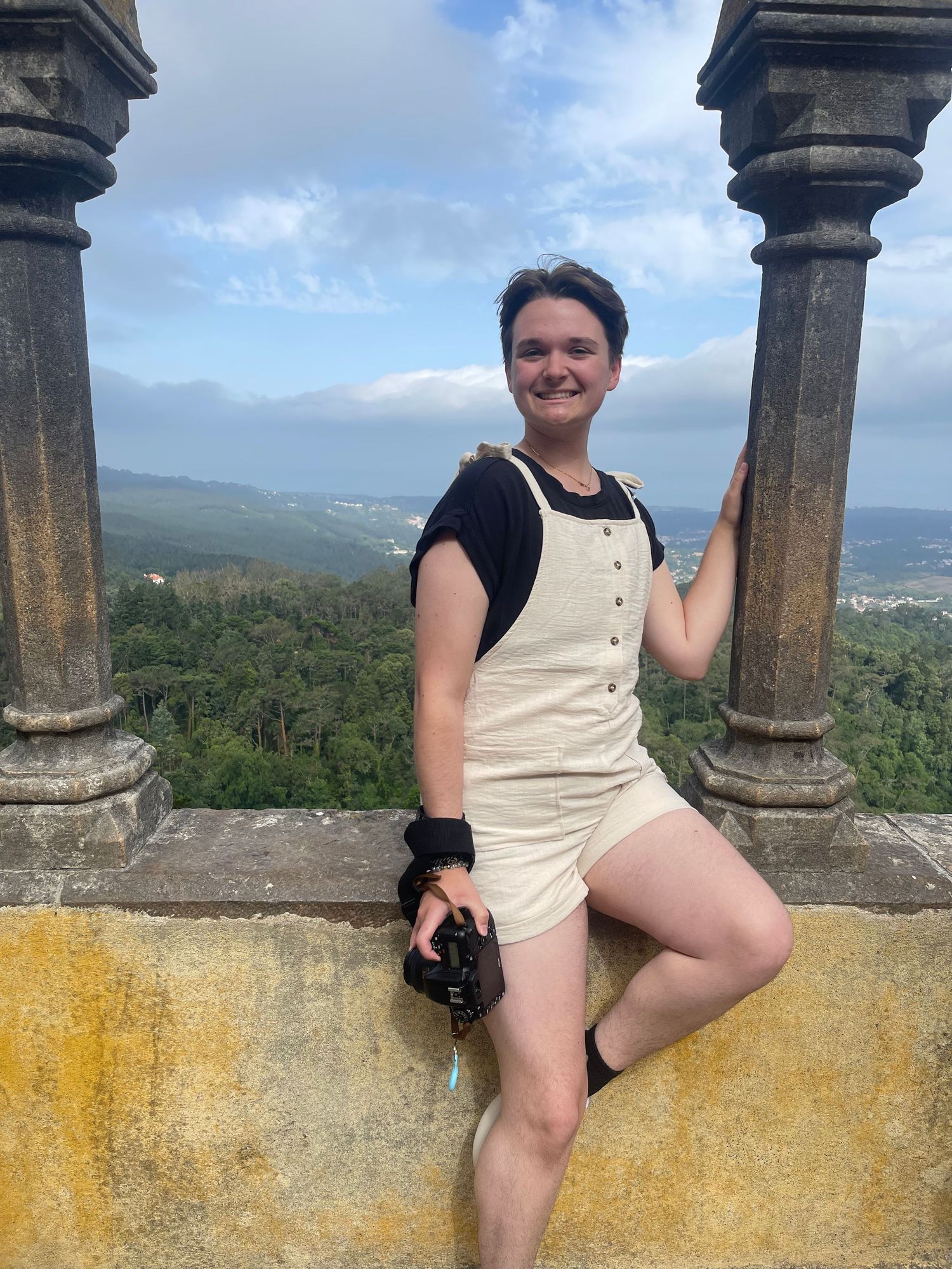 Someone poses along a stone balcony overlooking a scenic mountain landscape on a cloudy day.