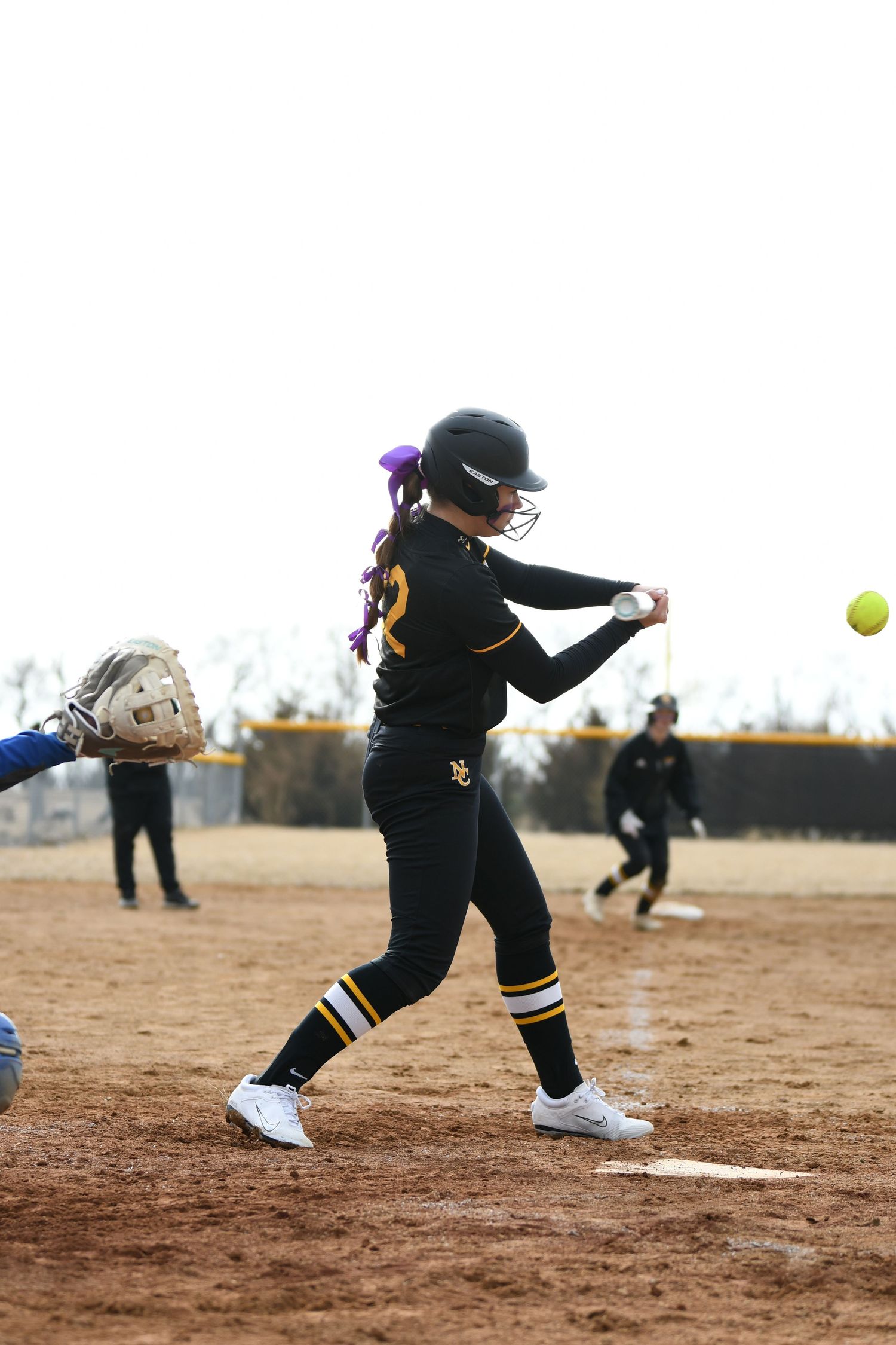 Softball batter in black uniform swinging at yellow ball on dirt field.
