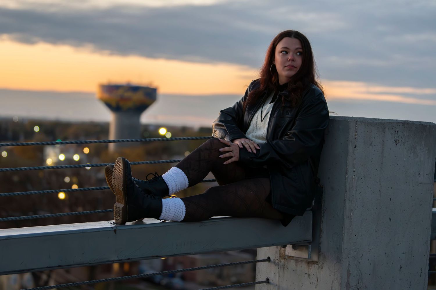 Person relaxing on rooftop ledge during sunset with city lights in background.