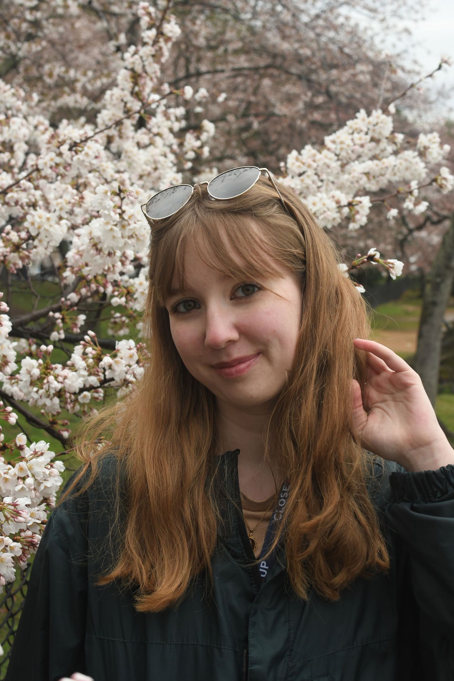 Person with sunglasses on head poses in front of blooming cherry blossom trees.
