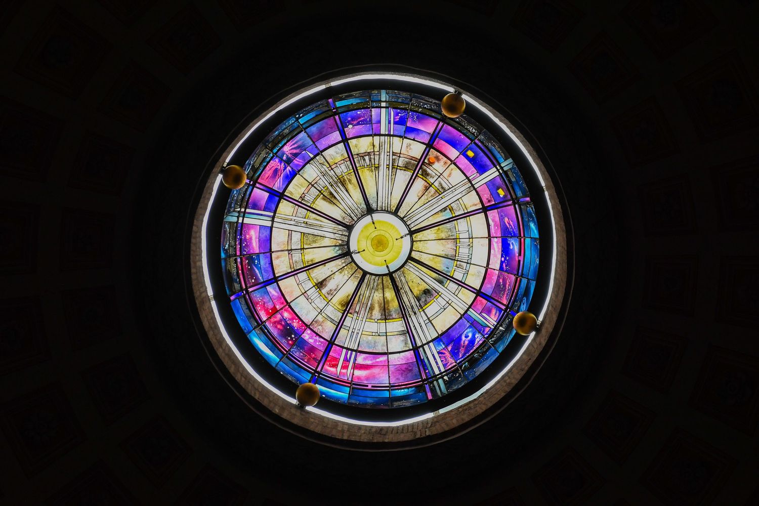 Colorful stained glass window with circular pattern in vibrant purple, blue and yellow hues.