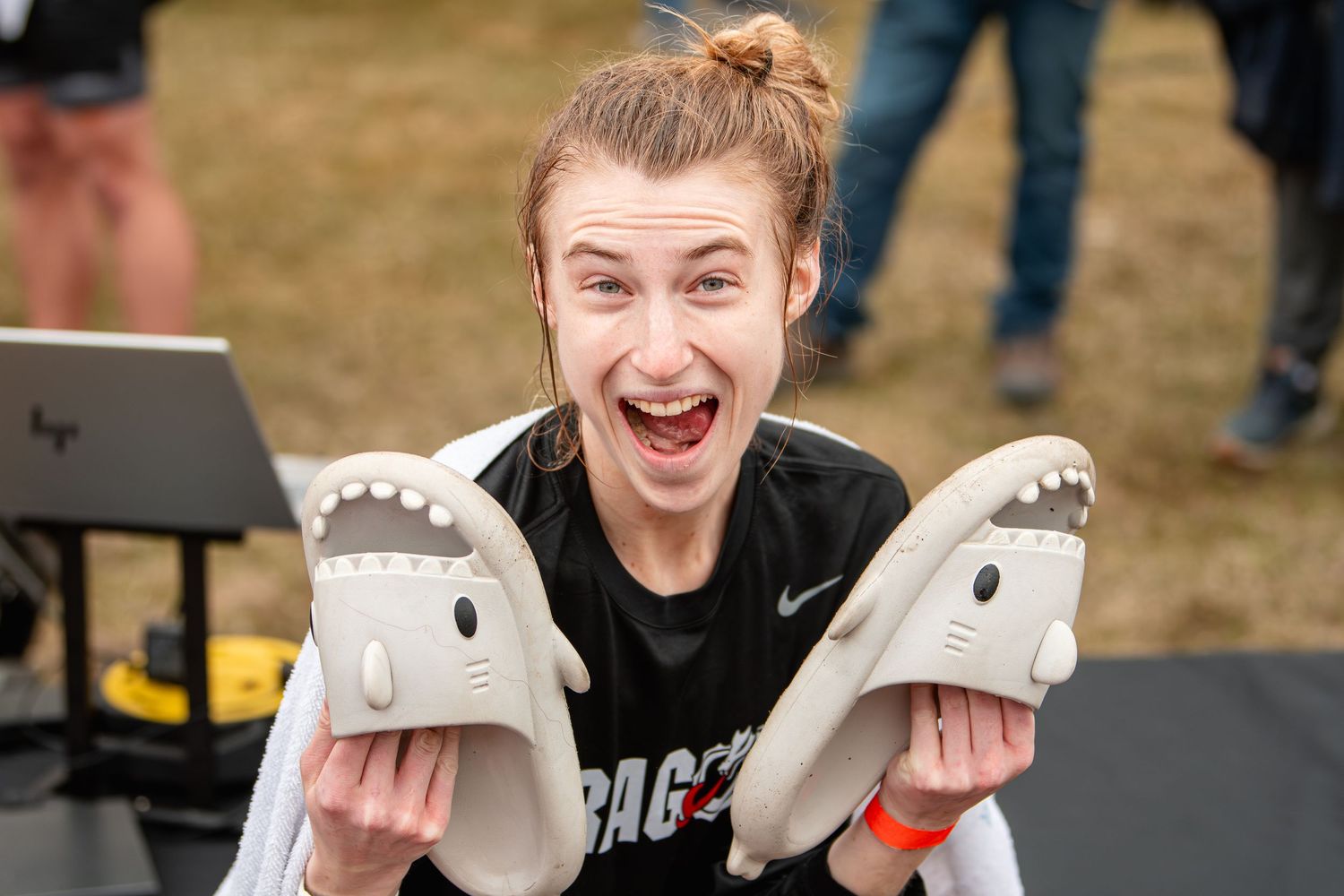 Excited young athlete holds shark-shaped slippers while wearing a black Gonzaga shirt outdoors at a sporting event.