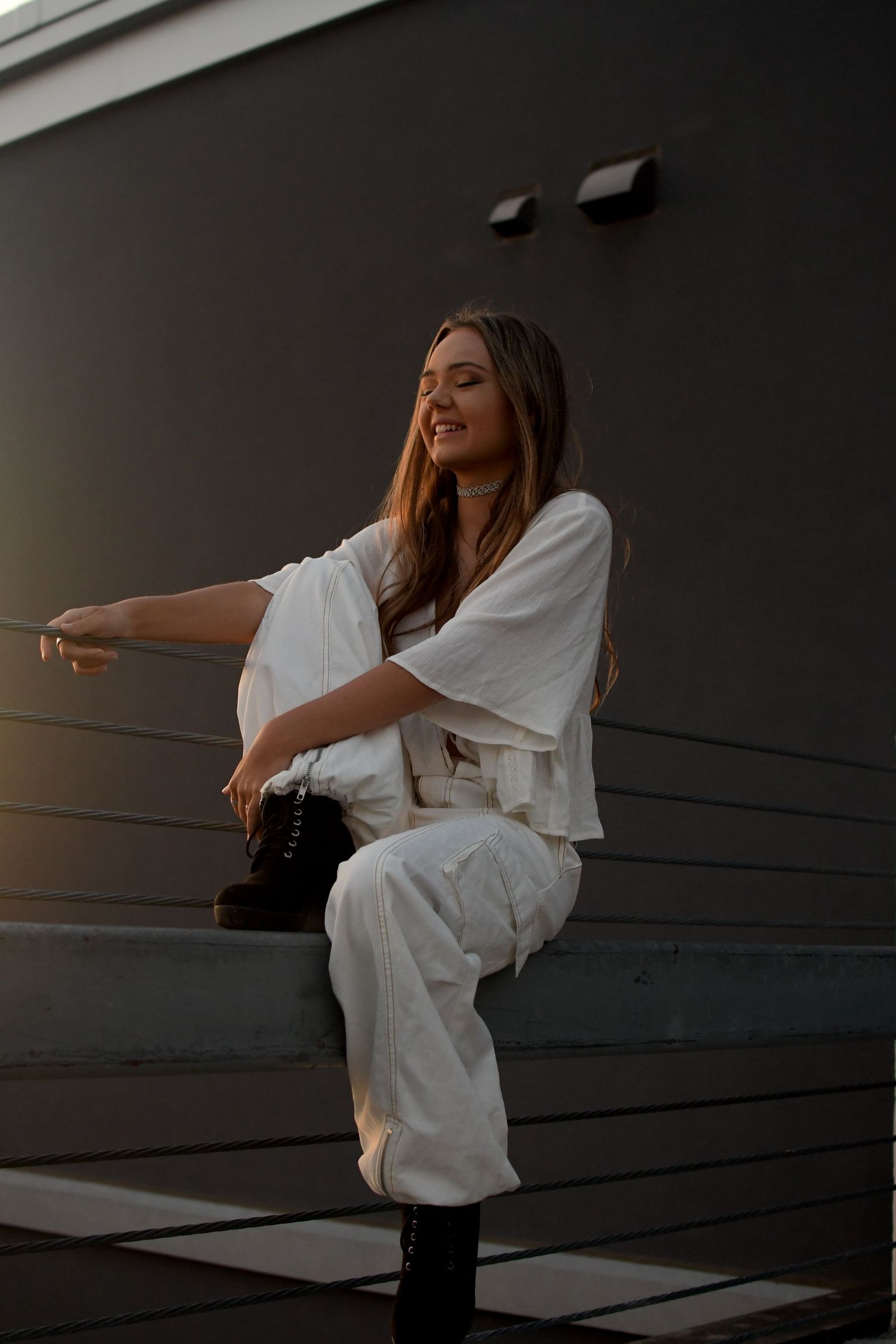 A person in white clothing sits on dark railing during sunset in an architectural setting creating a moody silhouette.