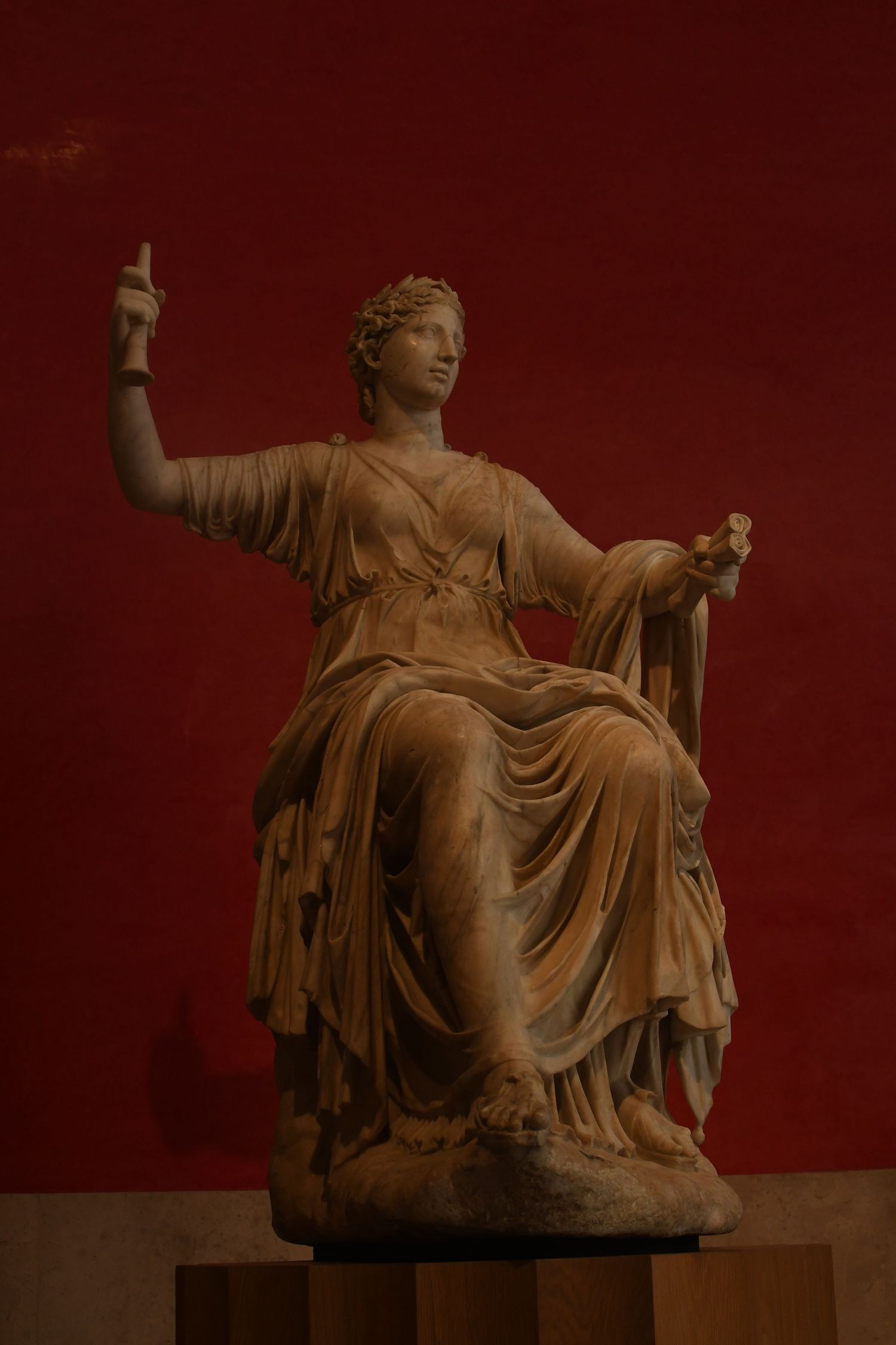 Classical Roman statue of a seated woman in draped robes against a deep red background.