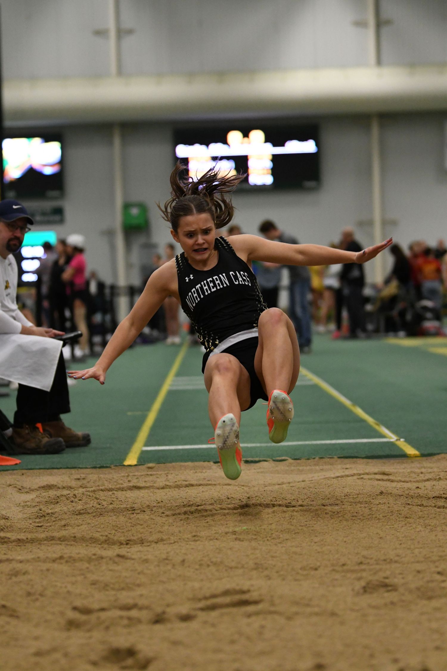 A long jumper soars through the air during an indoor track meet landing sequence.