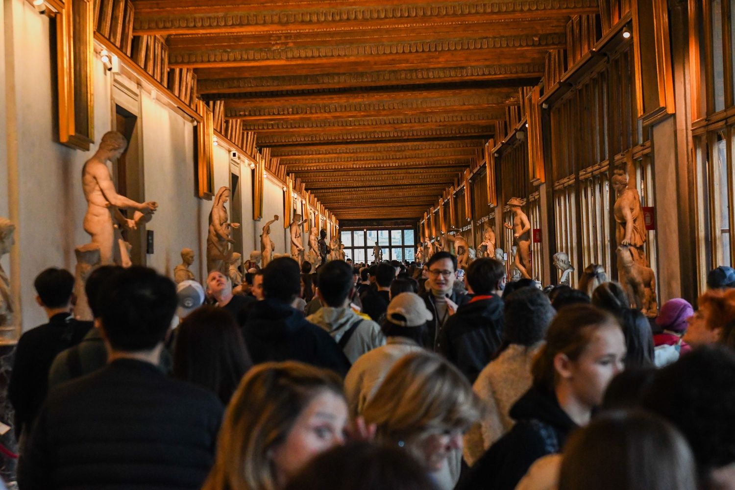 Crowded corridor in the Uffizi Gallery in Florence with wooden ceiling and artwork displays.