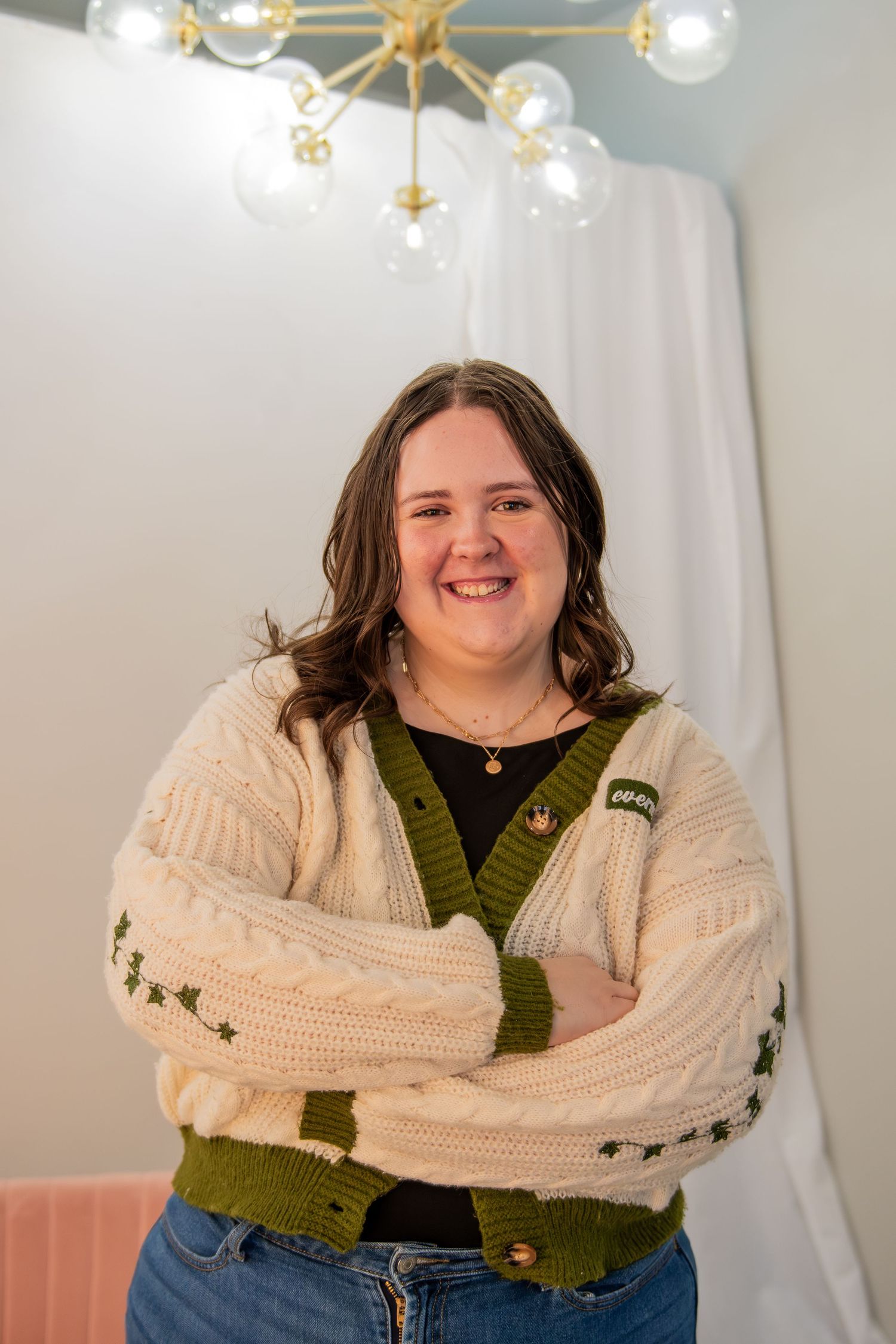 A person wearing a cream-colored knit sweater and olive green cardigan poses in front of white curtains and lights.