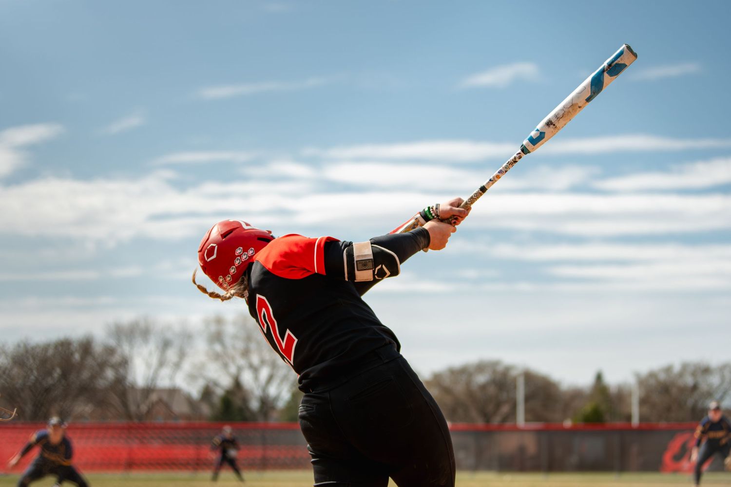Softball player in black and red uniform swinging bat during outdoor game under blue sky.