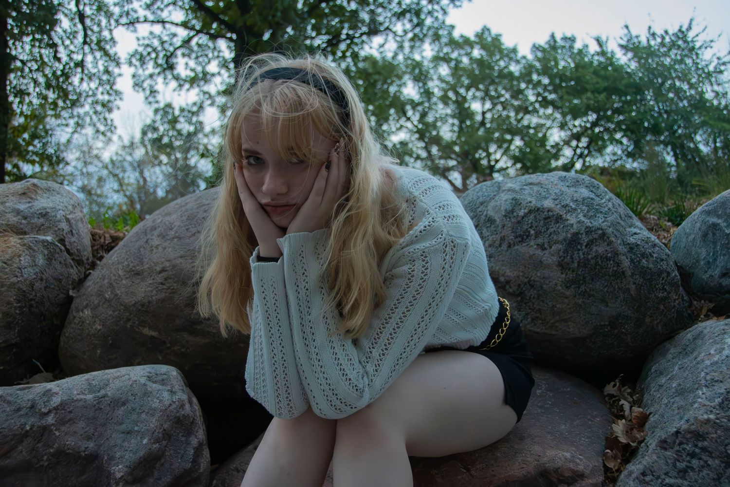 A person in a cream knit sweater poses thoughtfully on large rocks in an outdoor setting during autumn.